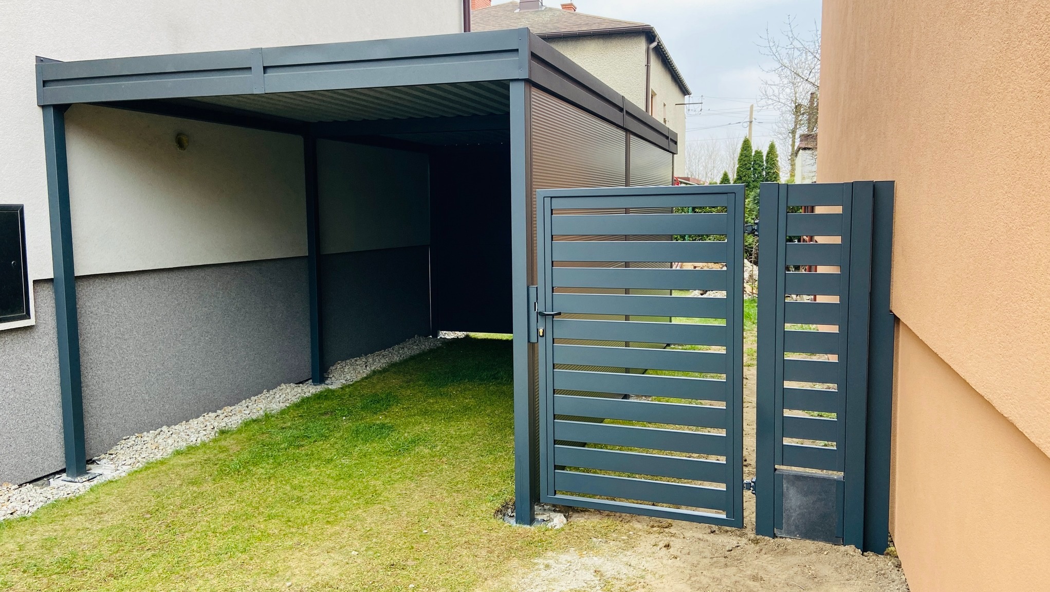Nowoczesna, grafitowa konstrukcja z metalu: zadaszenie i brama lamelowa na tle zielonego trawnika i elewacji budynku. Minimalistyczny design.