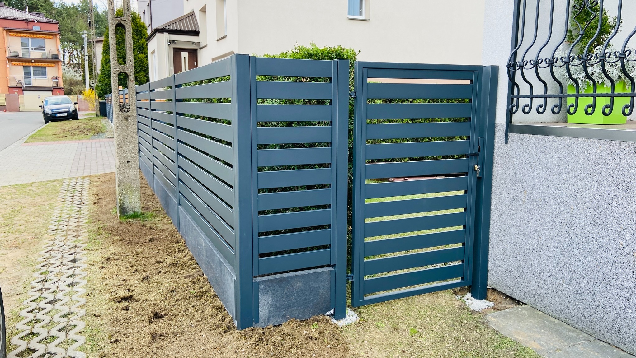 Nowoczesne, metalowe ogrodzenie panelowe w kolorze grafitowym z furtką, na tle zieleni i zabudowy jednorodzinnej. Minimalistyczny design, poziome lamele.