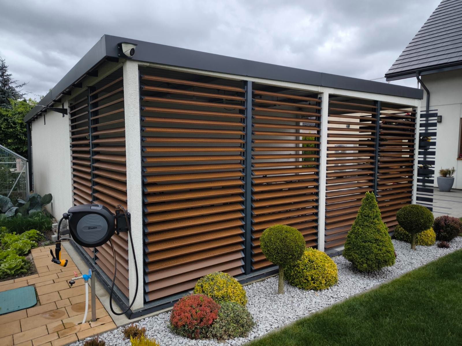 Nowoczesna, zamknięta pergola z drewnianymi żaluzjami, otoczona dekoracyjnymi krzewami i białym żwirem. Widoczny zwijany wąż ogrodowy i kamera na elewacji.