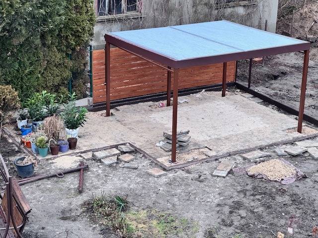 Nowoczesna, brązowa pergola metalowa z przezroczystym zadaszeniem na przygotowanym podłożu z kostki i piasku w ogrodzie.