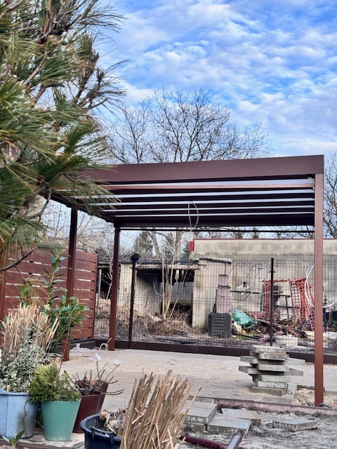 Metalowa pergola w kolorze brązowym z poziomymi lamelami, widoczna z boku w ogrodzie. Tło stanowi zabudowa z siatki i roślinność.