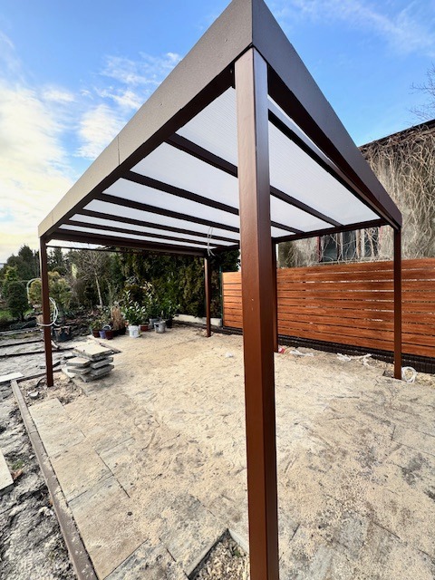 Nowoczesna, brązowa pergola metalowa z przezroczystym zadaszeniem na posesji, widok z dołu, elementy montażowe na ziemi.
