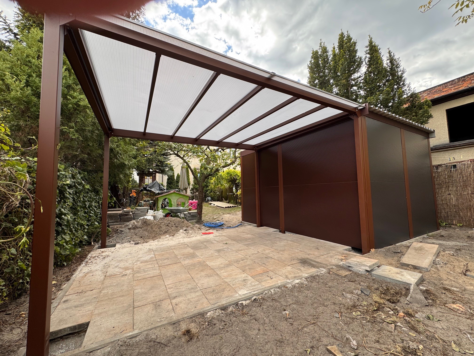 Brązowa, metalowa konstrukcja z białym zadaszeniem, częściowo zabudowana, na wyłożonym kostką placu. Widoczne elementy ogrodu i budynku w tle.