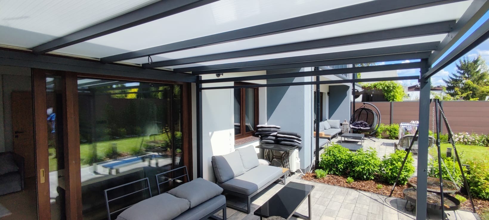Zadaszona metalowa konstrukcja tarasu z meblami ogrodowymi i roślinami. Widok na ogród przez przesuwne drzwi. Nowoczesny design i komfortowa przestrzeń relaksu.