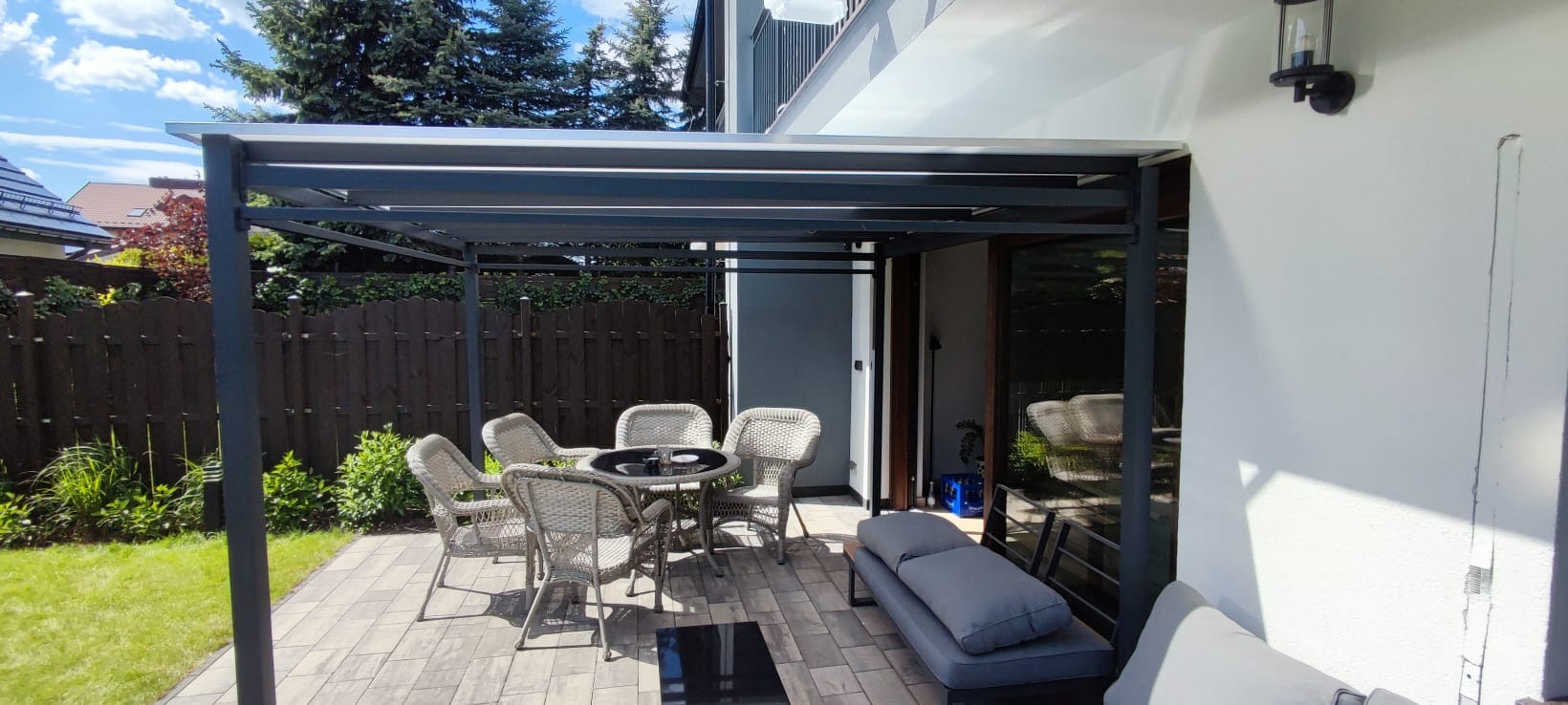 Nowoczesna, grafitowa pergola metalowa z meblami ogrodowymi na tarasie z kostki, przylegająca do jasnego budynku. Widok na zadbany ogród i drewniany płot.