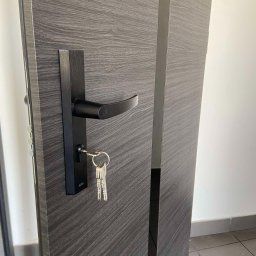 MAT-BUD Andrzej Winiarski - Ciemnoszare drzwi wejściowe z czarną klamką i kompletem kluczy w zamku, widoczne usłojenie drewna i metalowe wykończenia.