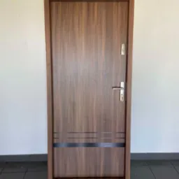 Brązowe drzwi wewnętrzne z metalową klamką i poziomymi, ciemnymi paskami dekoracyjnymi, ustawione na ciemnej podłodze na tle białej ściany.