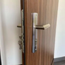 Brązowe drzwi z widocznym zamkiem wielopunktowym i chromowaną klamką, klucze w zamku, widoczna tekstura drewna.