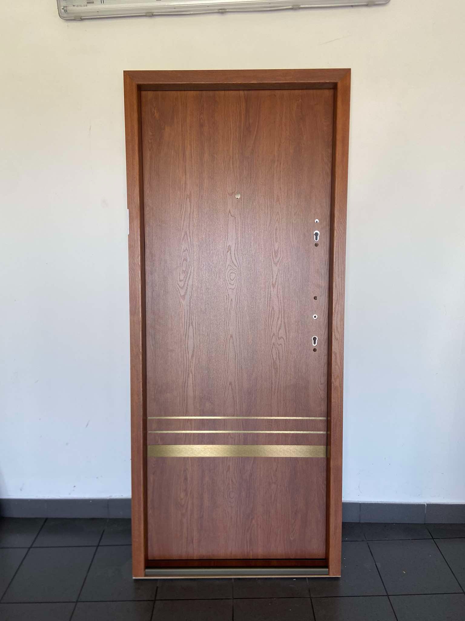 Brązowe drzwi antywłamaniowe z trzema złotymi paskami dekoracyjnymi, widoczne zamki i wizjer, na tle białej ściany i ciemnej podłogi z płytek.
