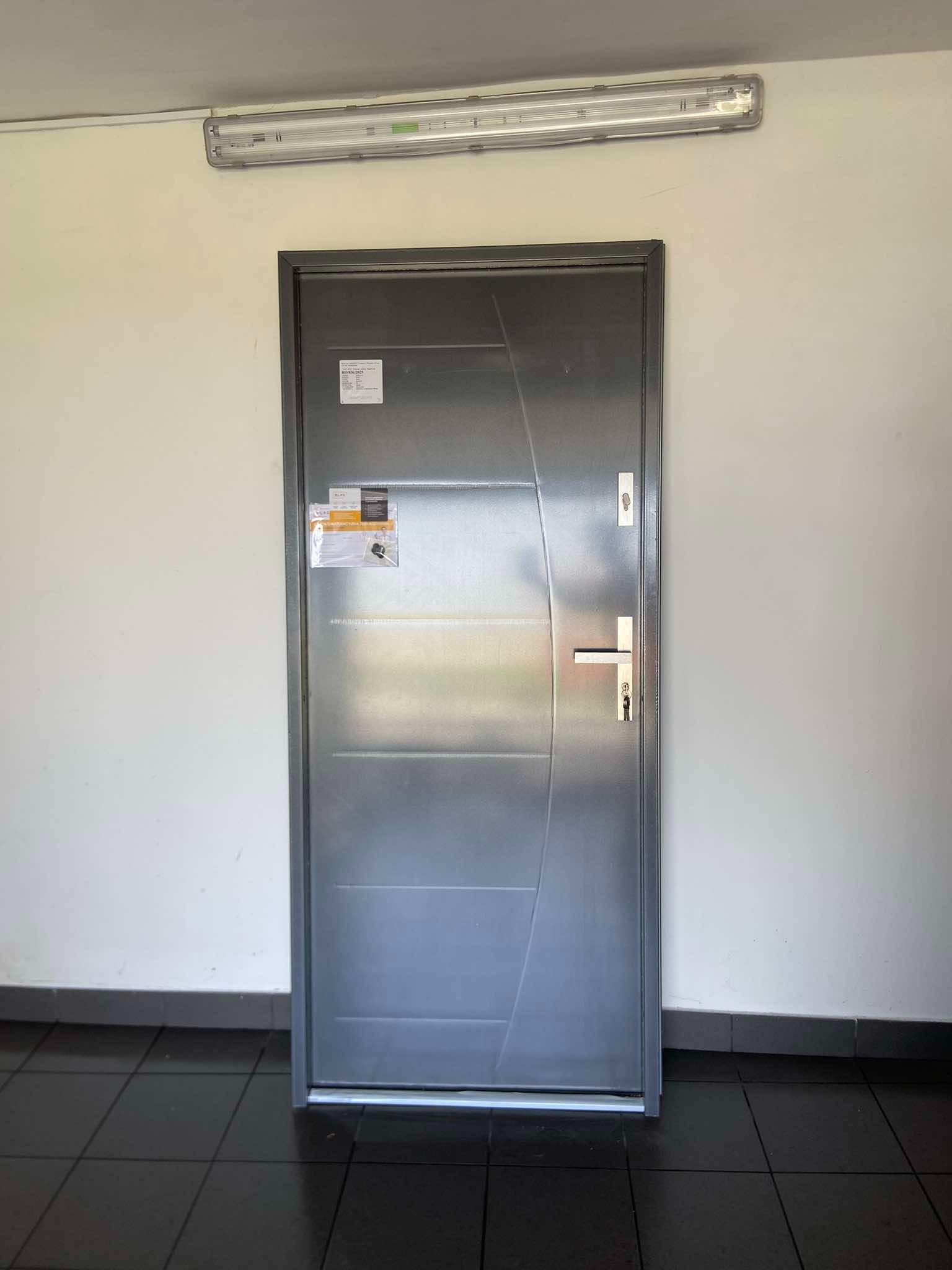 Szare drzwi wejściowe z naklejkami informacyjnymi, zamontowane w korytarzu z czarną podłogą i oświetleniem jarzeniowym.