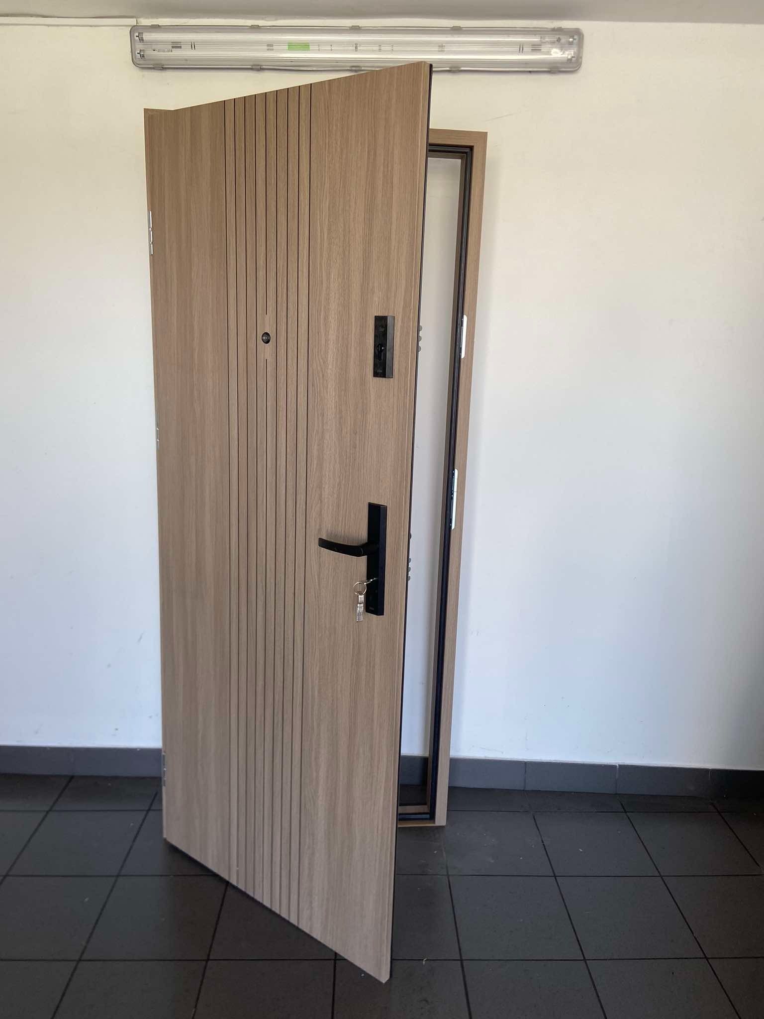 Otwarta, nowoczesna drzwi wejściowe z panelem w odcieniach drewna i pionowymi frezowaniami, czarną klamką i zamkiem z kluczami, widoczna ościeżnica w ciemnym kolorze, na tle białej ściany i ciemnej...