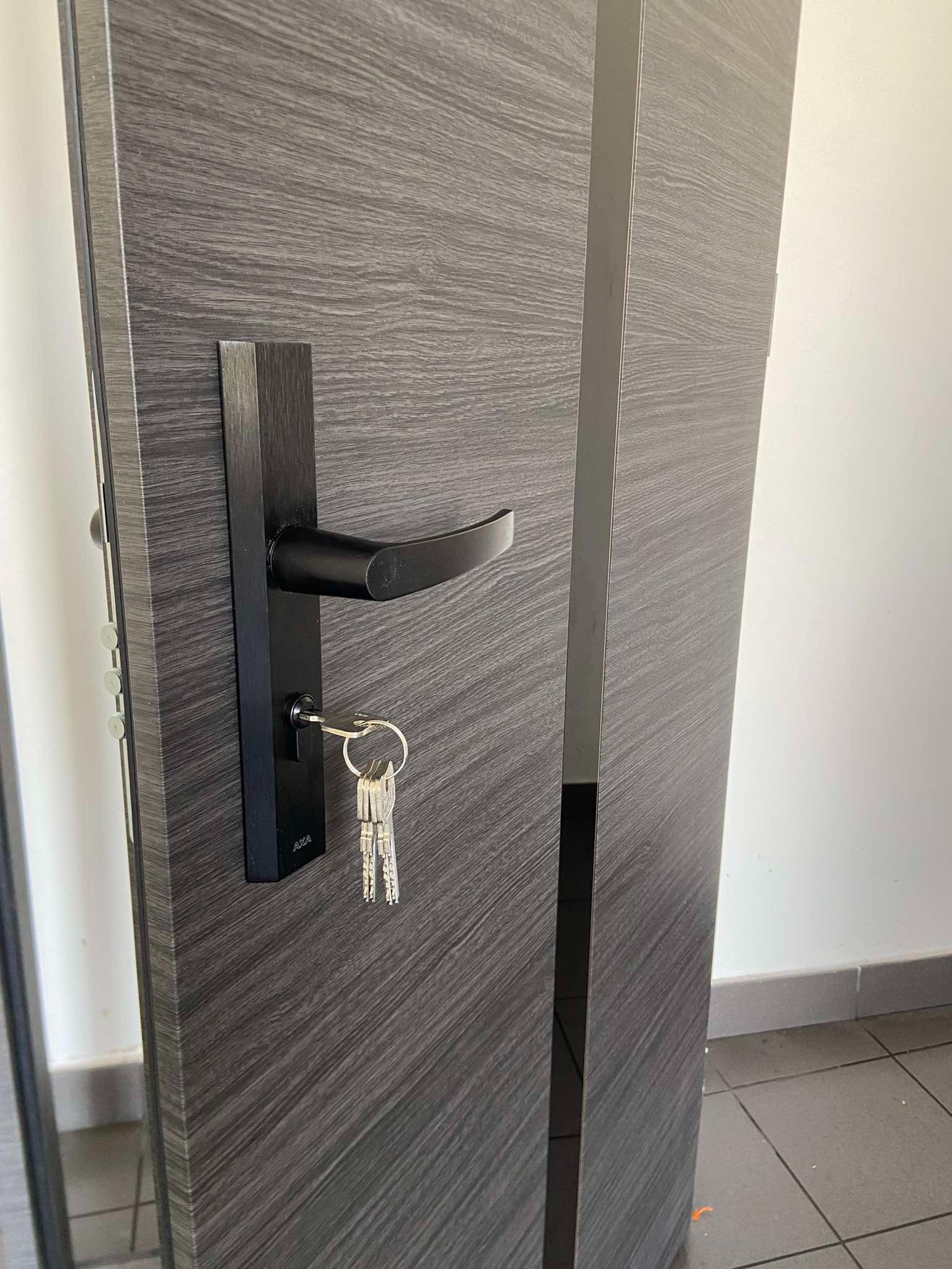 Ciemnoszare drzwi wejściowe z czarną klamką i kompletem kluczy w zamku, widoczne usłojenie drewna i metalowe wykończenia.