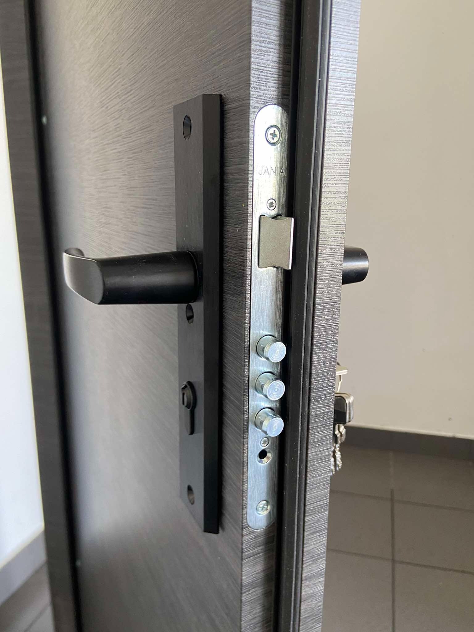 Częściowo otwarta, ciemna, nowoczesna brama wejściowa z widocznym wielopunktowym zamkiem listwowym marki Jana i czarną klamką, widoczne klucze w zamku.