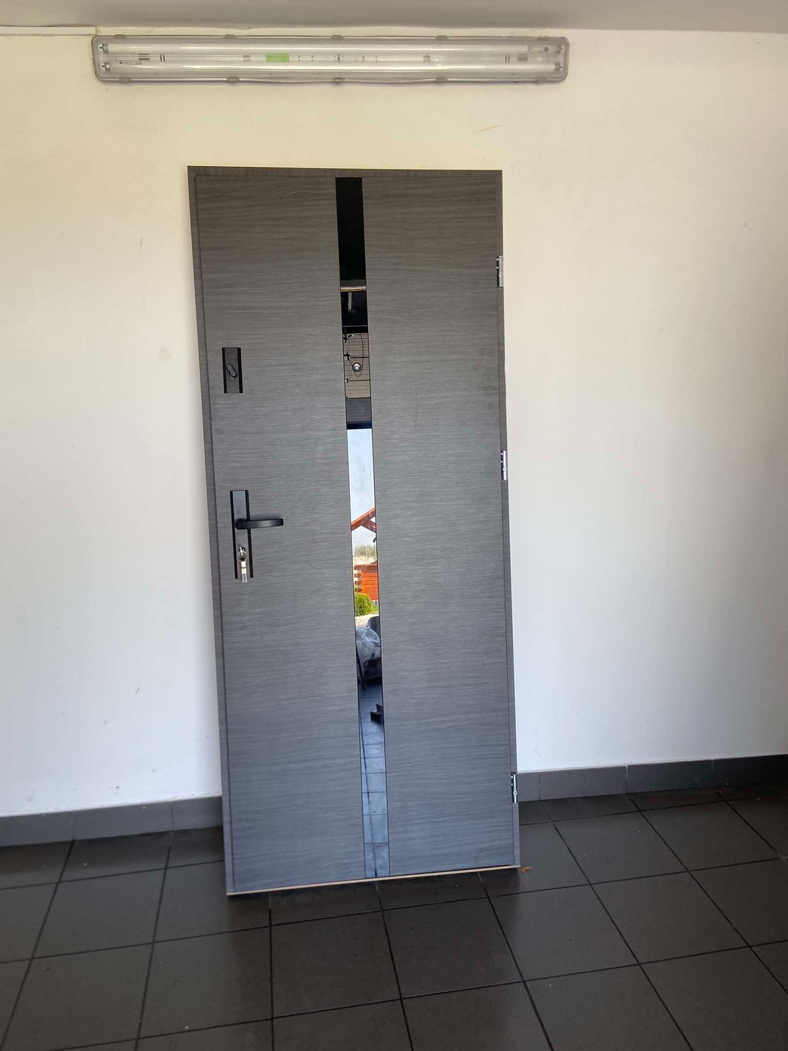 Szare drzwi wejściowe z pionowym pasem lustra pośrodku, widok z zewnątrz, zamek i klamka po lewej stronie, widoczne elementy montażowe, surowy korytarz z czarną podłogą w płytkach.