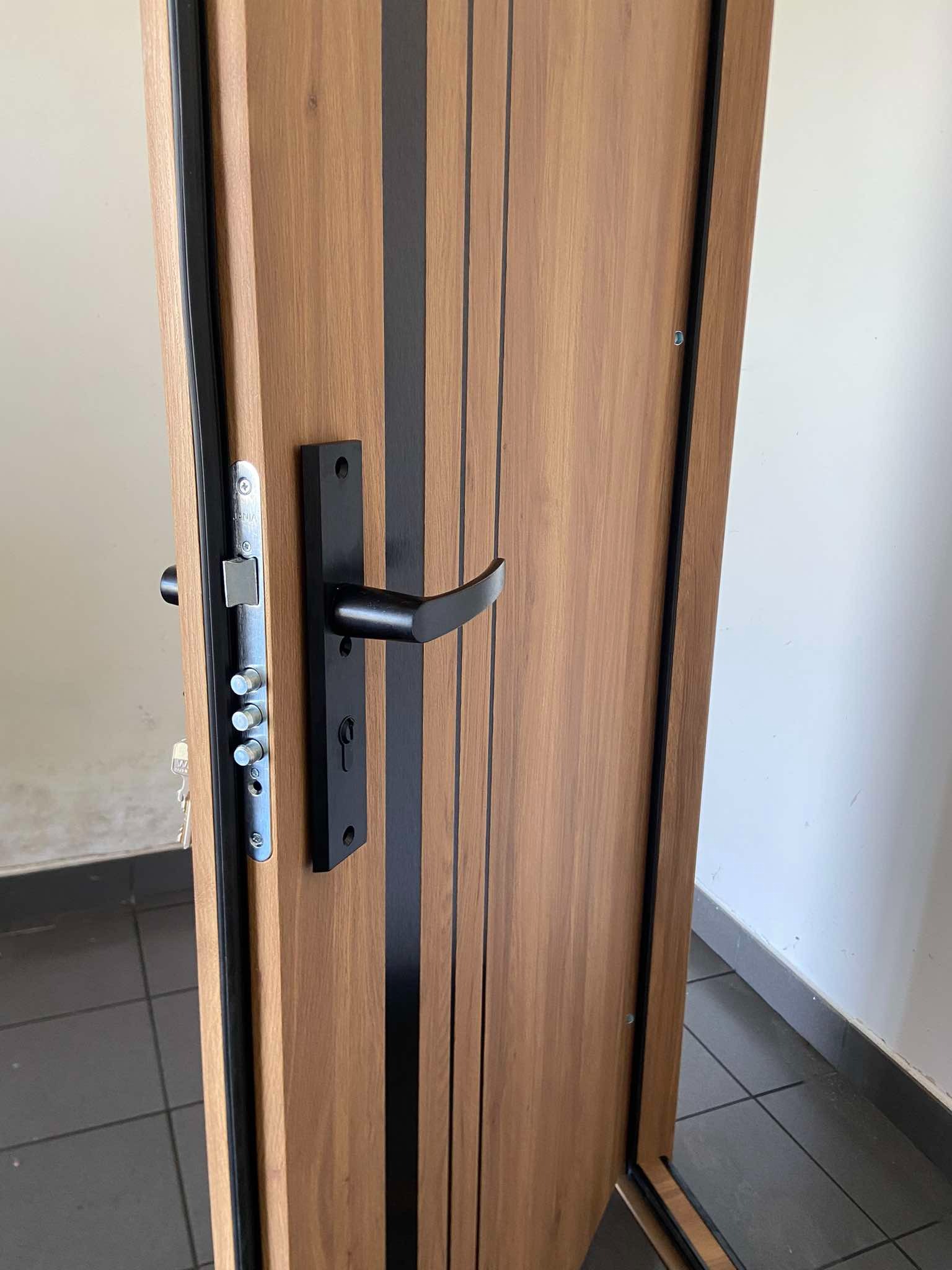 Otwarta, drewnopodobna drzwi antywłamaniowe z czarnymi akcentami, widoczne mechanizmy zamka i klamka.