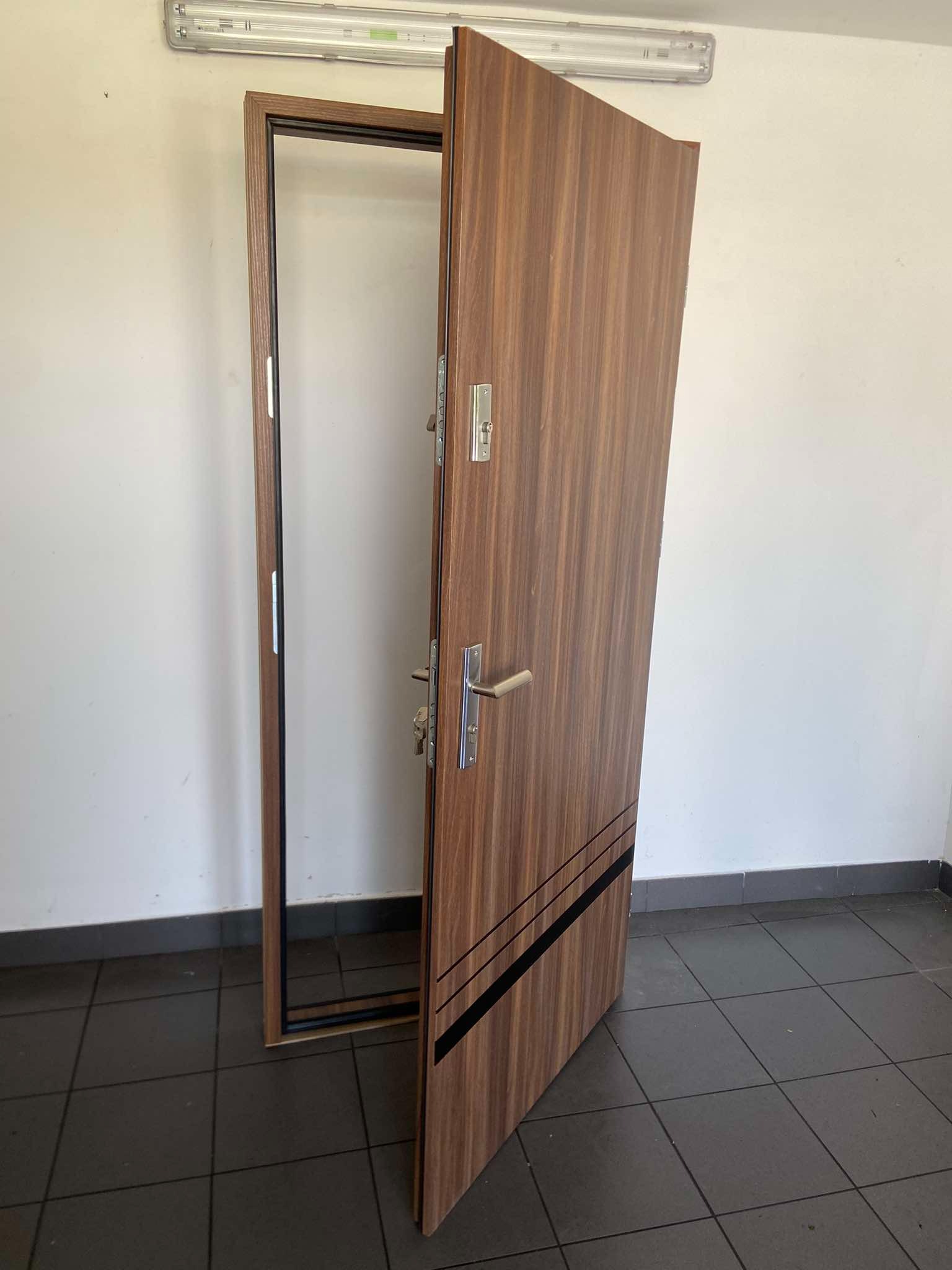 Otwarta, brązowa, drewnopodobna płyta drzwiowa z czarnymi paskami, zawieszona na ościeżnicy, widoczne zamki i klamka na tle białej ściany i ciemnej podłogi z płytek.