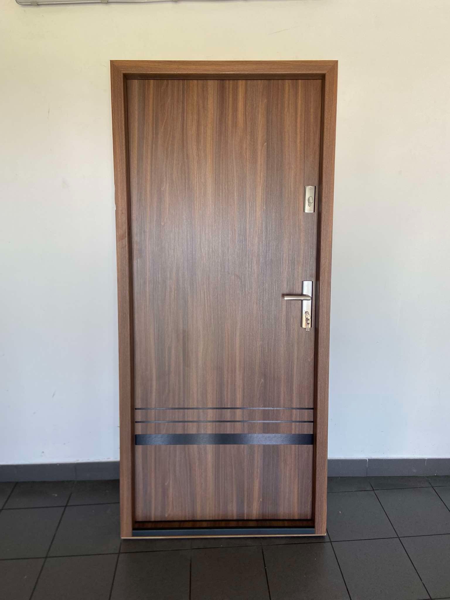 Brązowe drzwi wewnętrzne z metalową klamką i poziomymi, ciemnymi paskami dekoracyjnymi, ustawione na ciemnej podłodze na tle białej ściany.