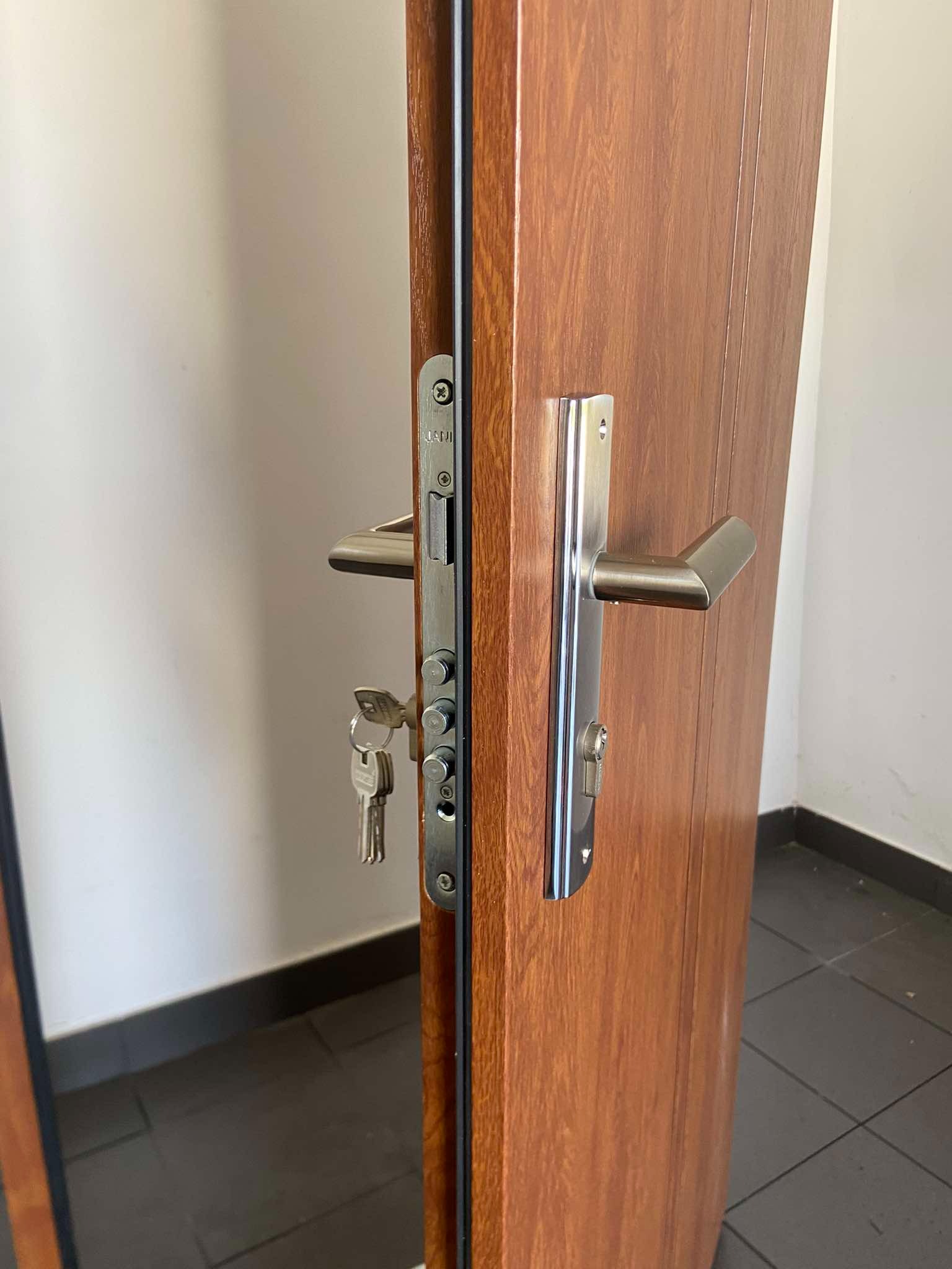 Brązowe drzwi z zamkiem wielopunktowym i kluczami w zamku, widoczne płytki podłogowe.