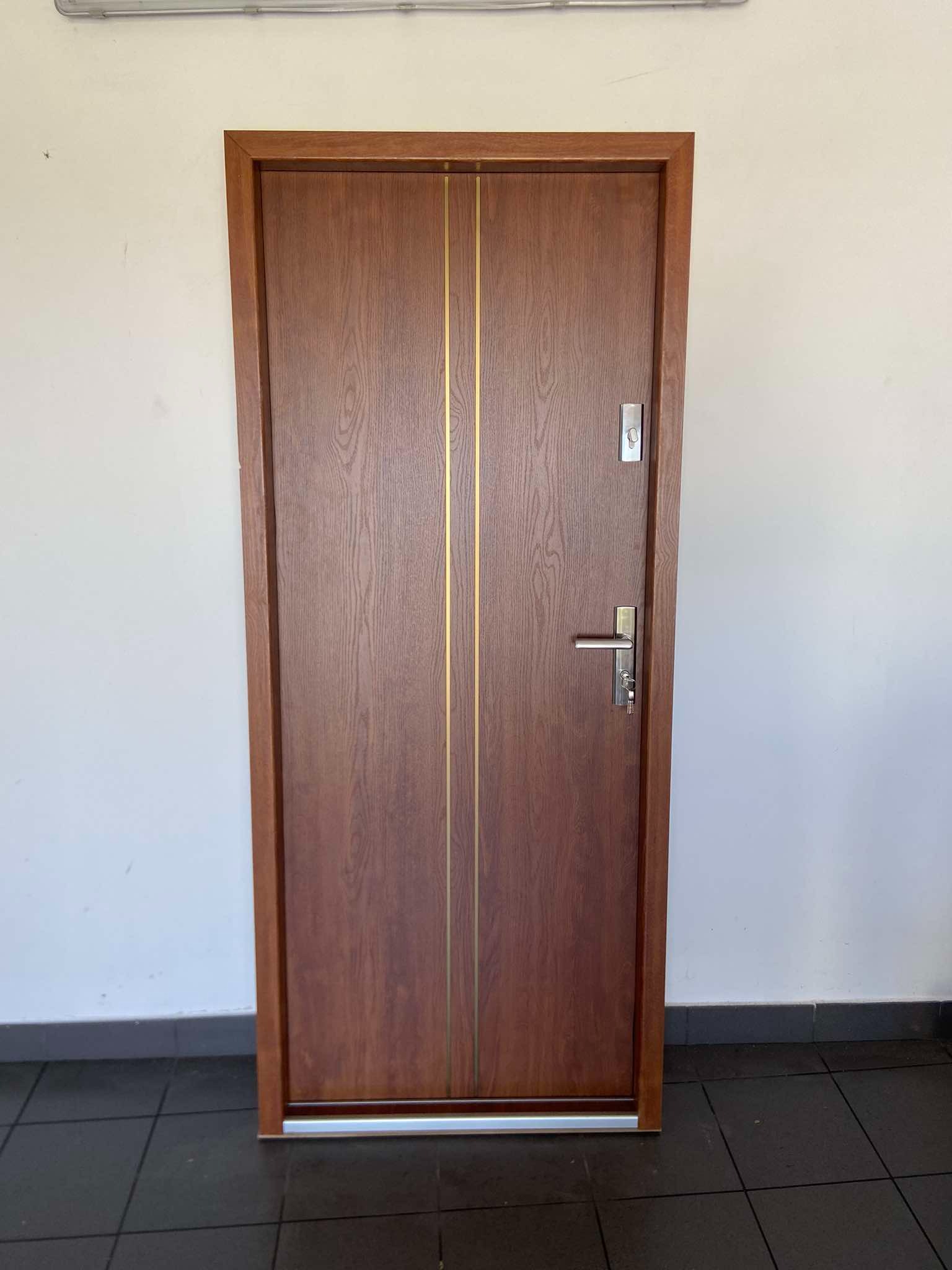Brązowe drzwi wejściowe z dwoma złotymi paskami, zamontowane w białej ścianie, widoczne klucze w zamku.