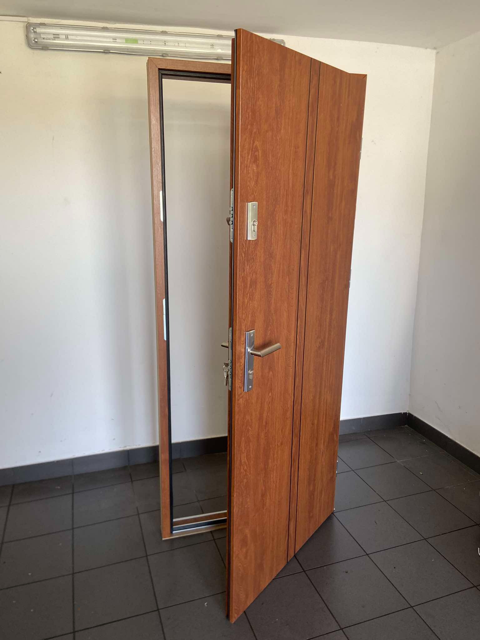 Otwarta, brązowa drzwi wejściowe z metalowymi elementami, widoczne zamki i klamka, na szarej podłodze z płytek, w pomieszczeniu z białymi ścianami i lampą na suficie.