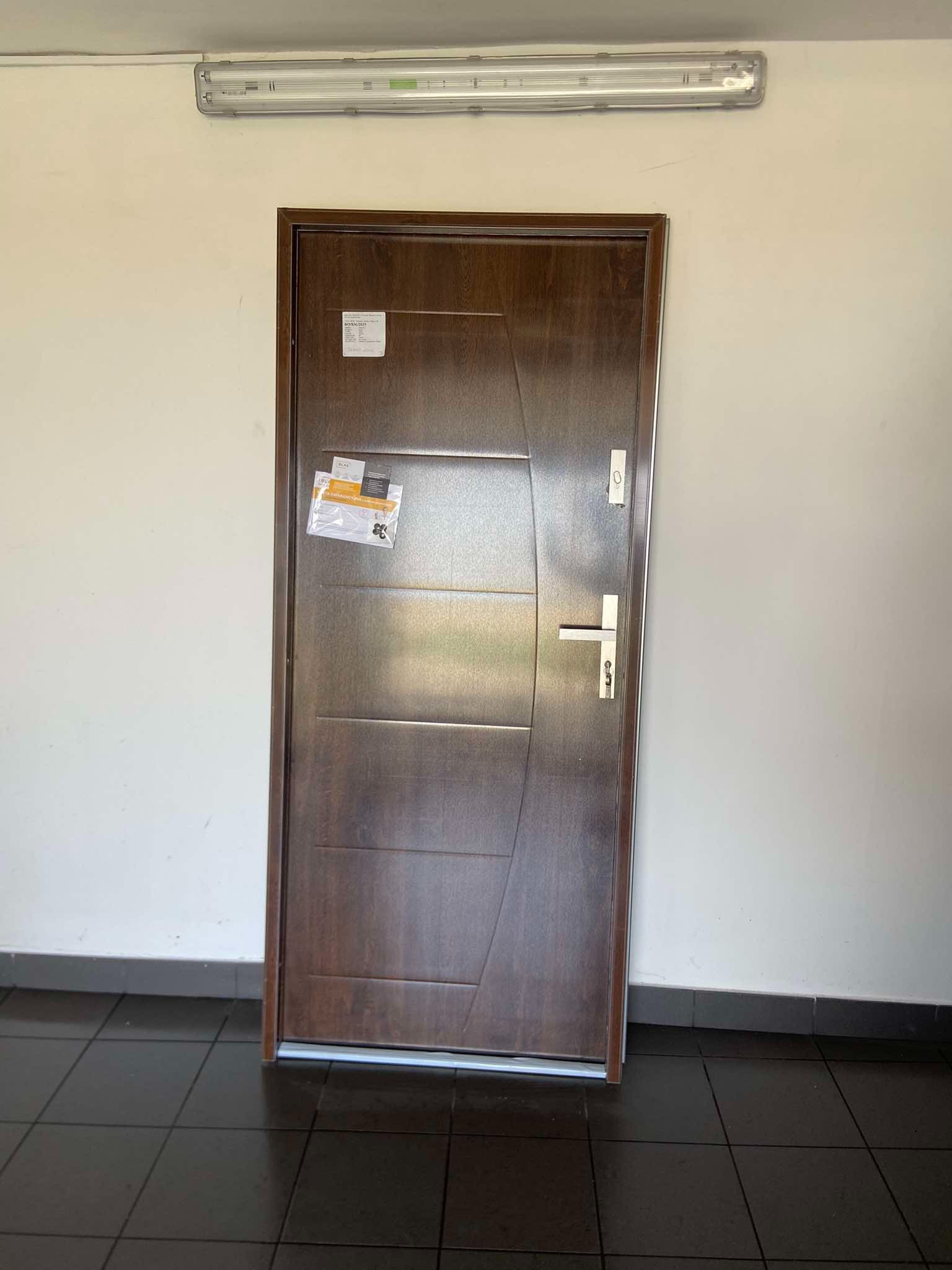 Ciemnobrązowe drzwi wejściowe z naklejkami, zamontowane w jasnym korytarzu z ciemną podłogą z płytek.