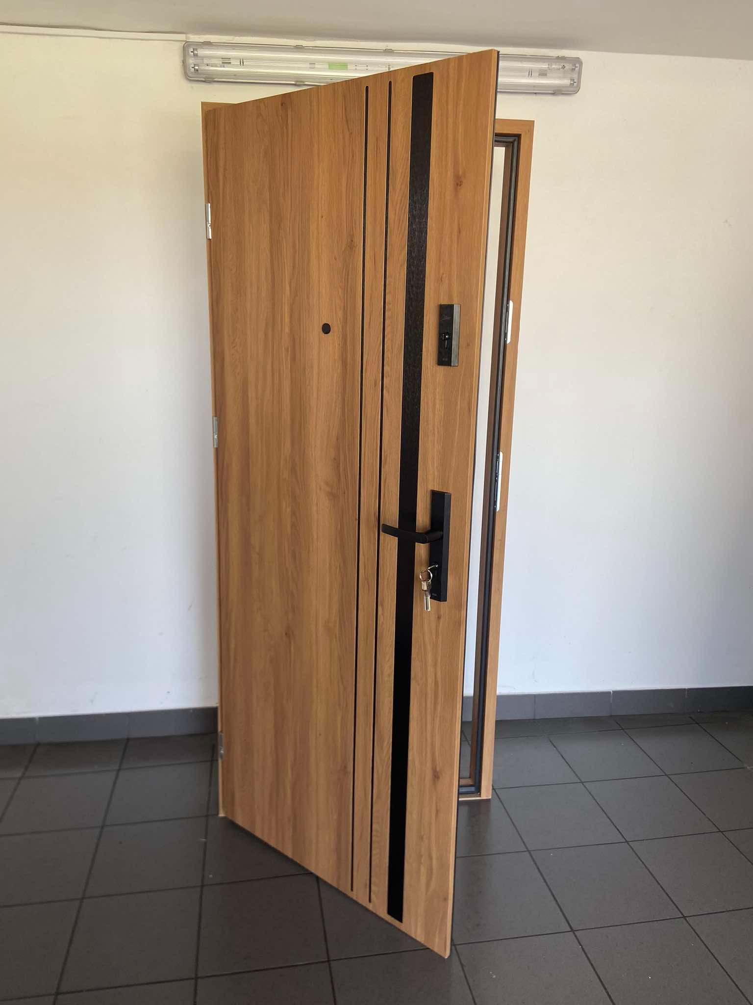 Otwarta drewniana drzwi antywłamaniowe z czarnymi wstawkami i wizjerem, widoczne klucze w zamku, na szarych płytkach podłogowych i białych ścianach, oświetlona jarzeniówką.