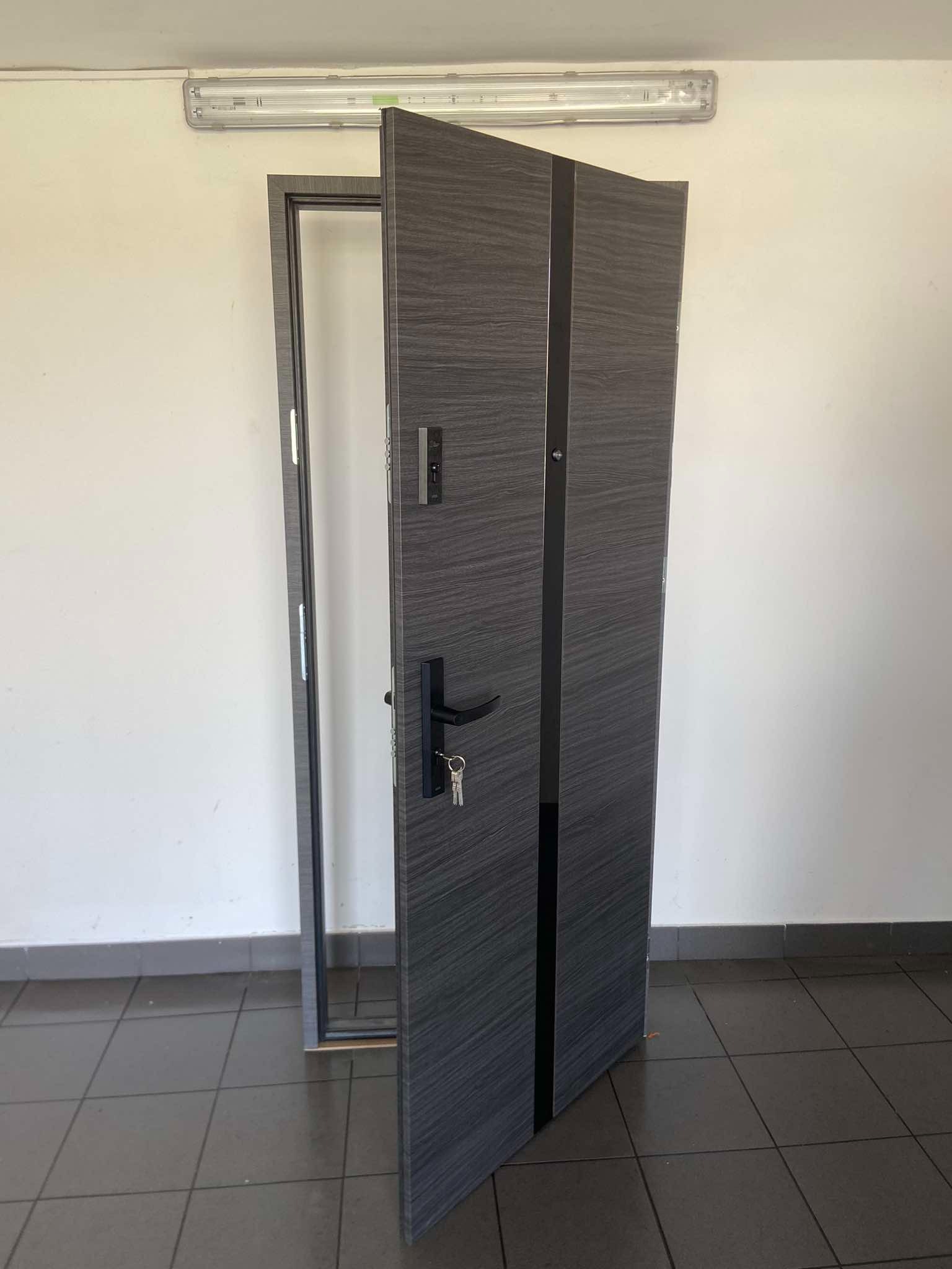 Częściowo zamontowane, nowoczesne drzwi wejściowe w odcieniach szarości z czarnymi akcentami, widoczne klucze w zamku, ościeżnica w trakcie montażu na tle białej ściany i szarej podłogi