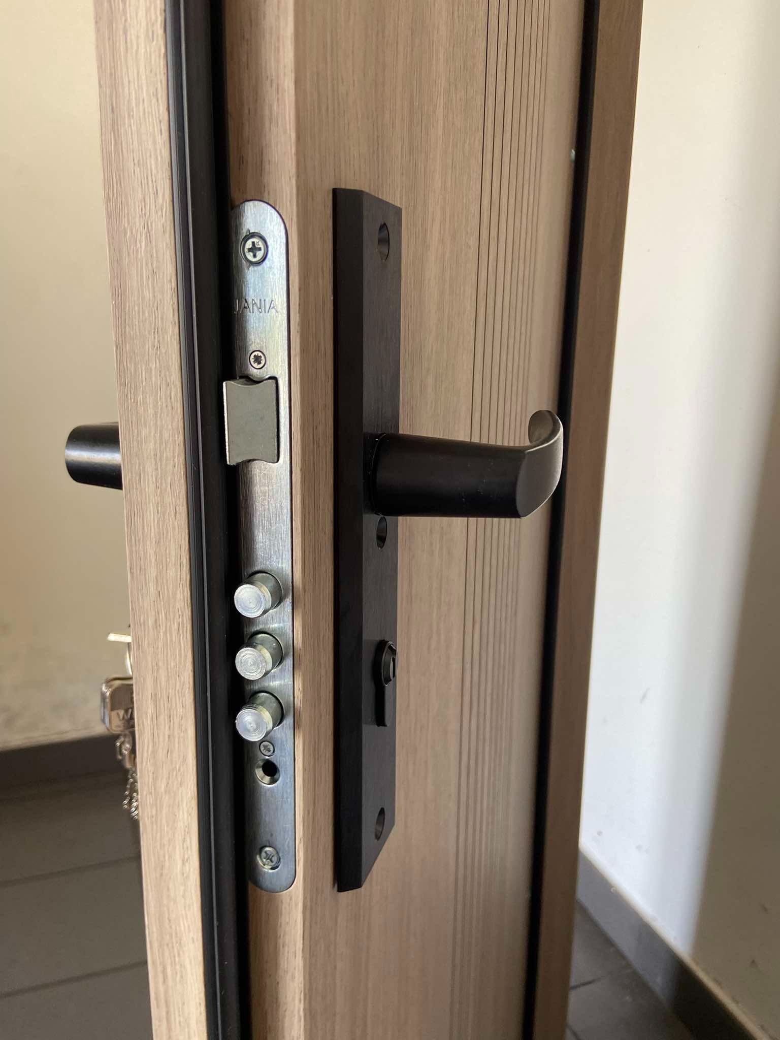 Zamek wielopunktowy i czarna klamka w drewnopodobnych drzwiach z pionowymi frezowaniami, widoczny fragment klucza.