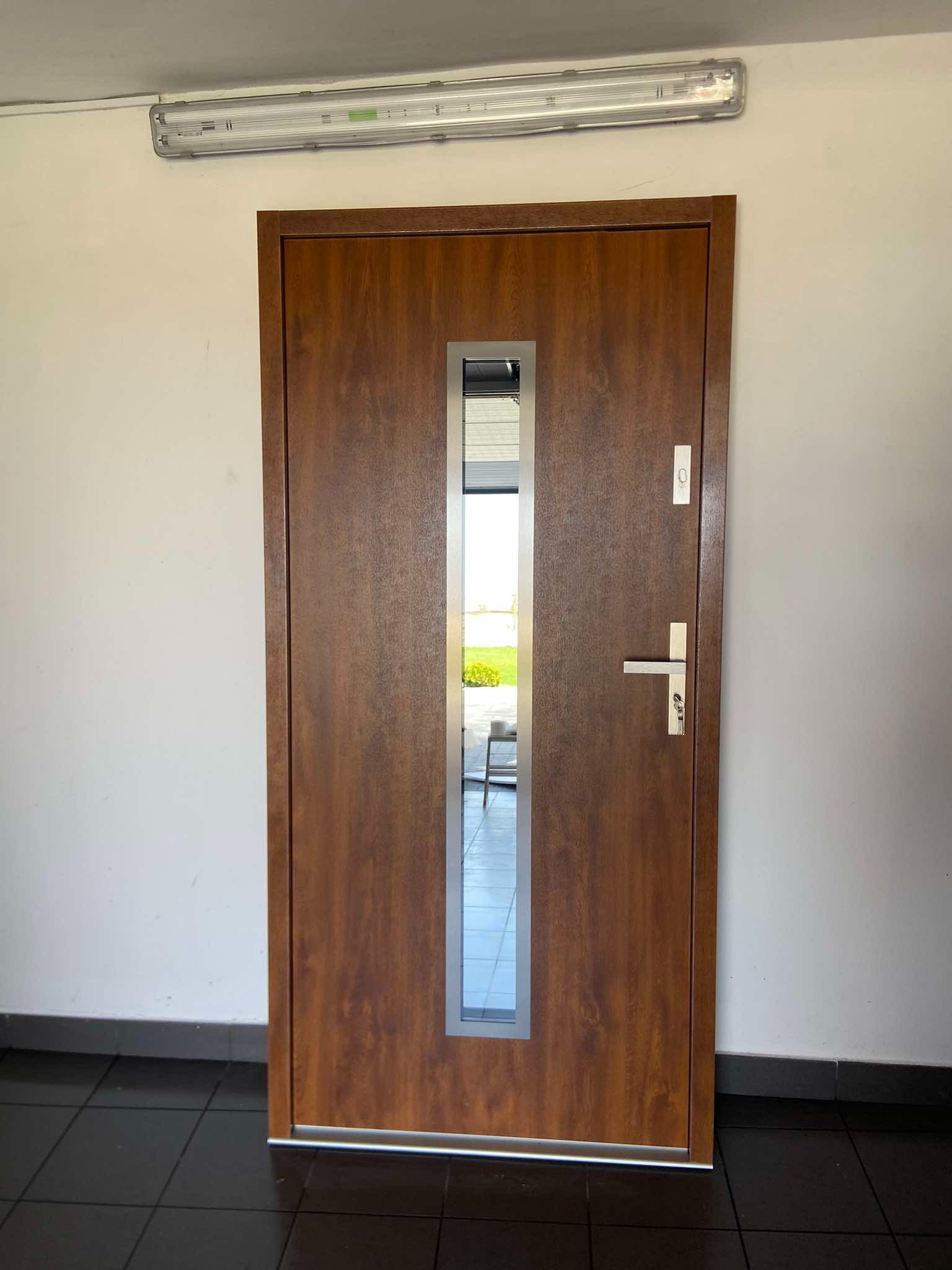 Brązowe drzwi wejściowe z wąskim przeszkleniem, widok na zewnątrz, srebrna klamka i wizjer na białej ścianie z czarną podłogą.