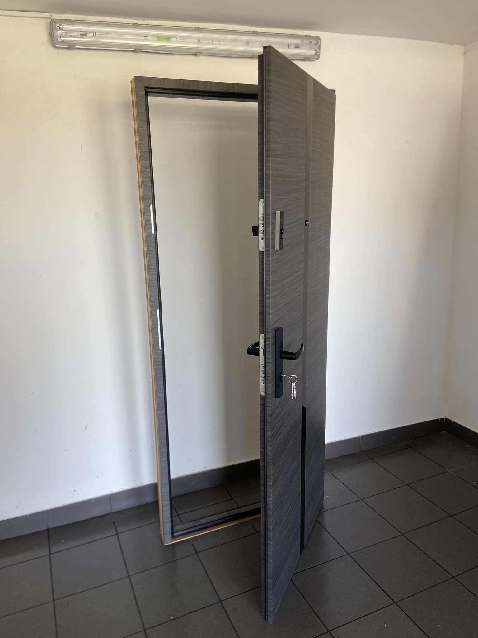 Otwarta, szara, zewnętrzna płyta drzwiowa z zamontowanym czarnym uchwytem i kluczem w zamku, widoczna rama drzwiowa z elementami montażowymi, na tle białej ściany i podłogi z ciemnych płytek.