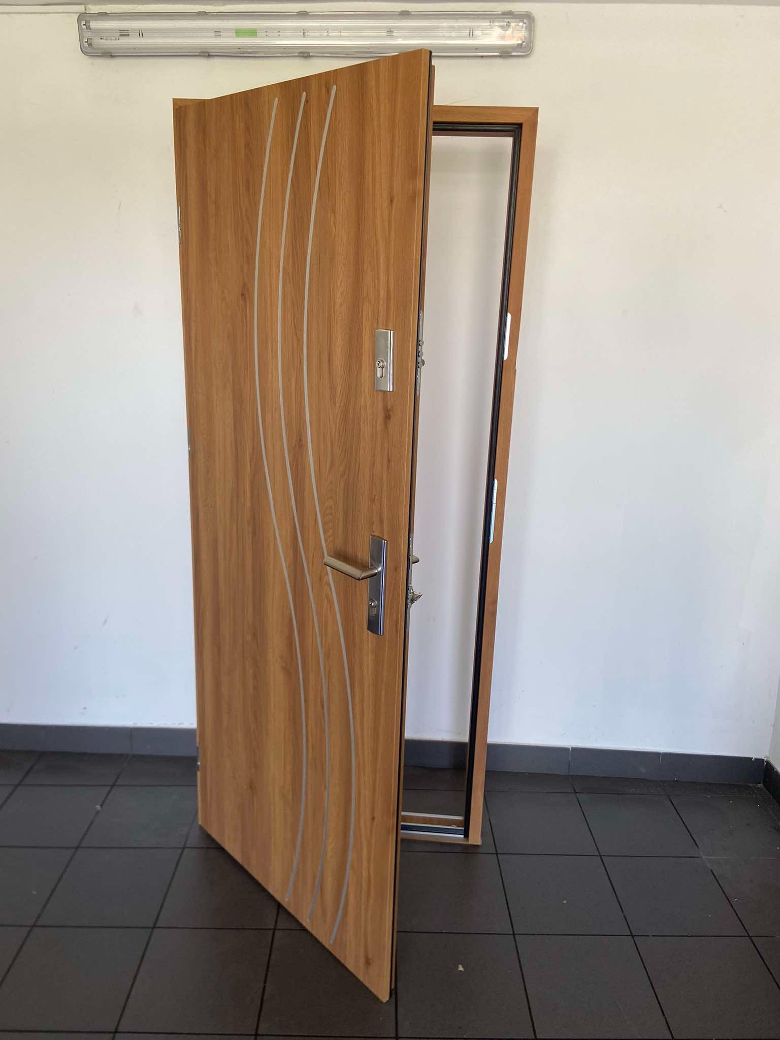 Otwarta, drewnopodobna, zewnętrzna płyta drzwiowa z trzema pionowymi, srebrnymi intarsjami i stalową klamką, widoczna ościeżnica z czarnym uszczelnieniem, szara ściana i ciemna podłoga...