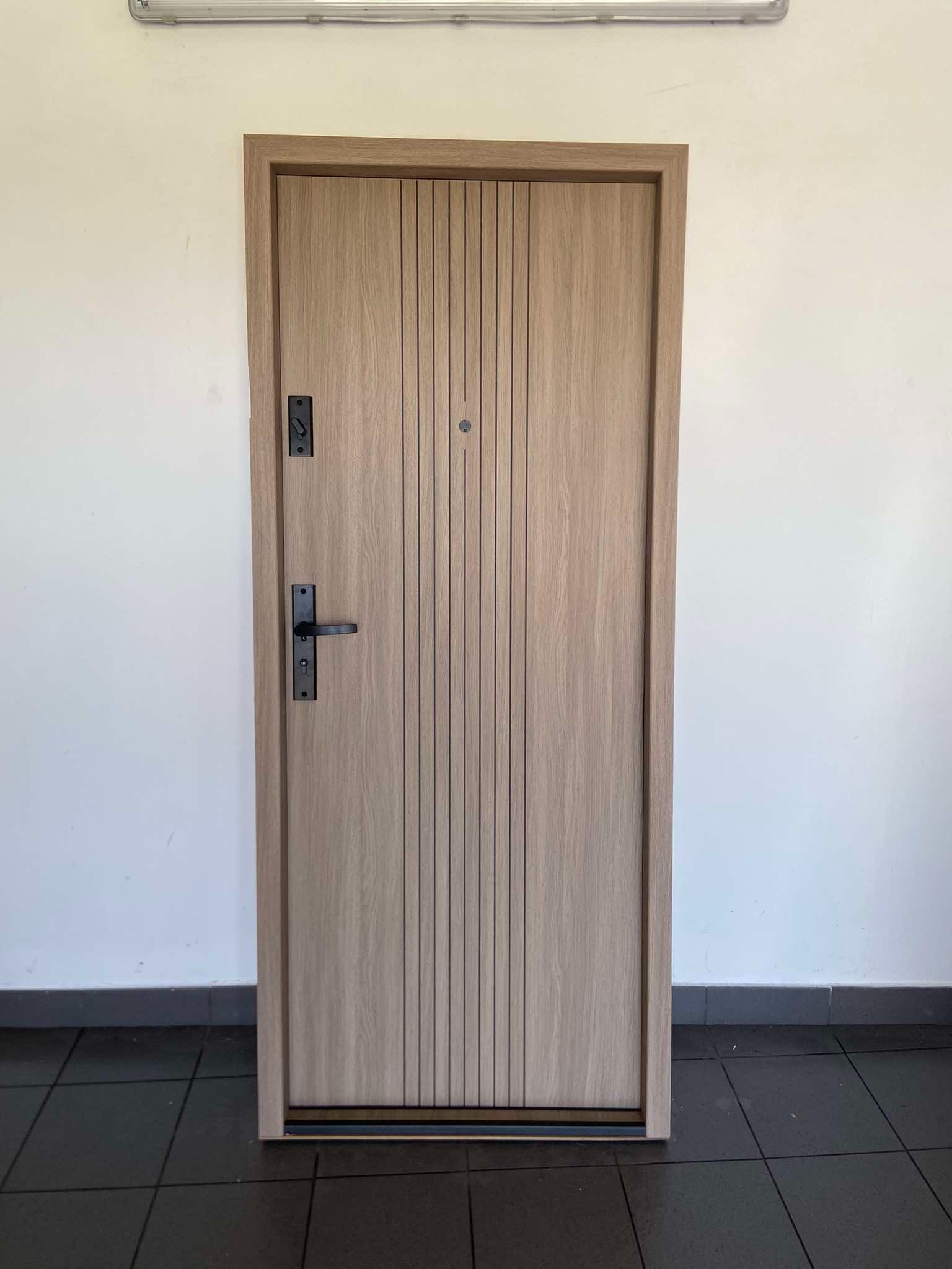 Jasne, drewnopodobne drzwi antywłamaniowe z pionowymi frezowaniami i czarną klamką na tle białej ściany i ciemnej podłogi.