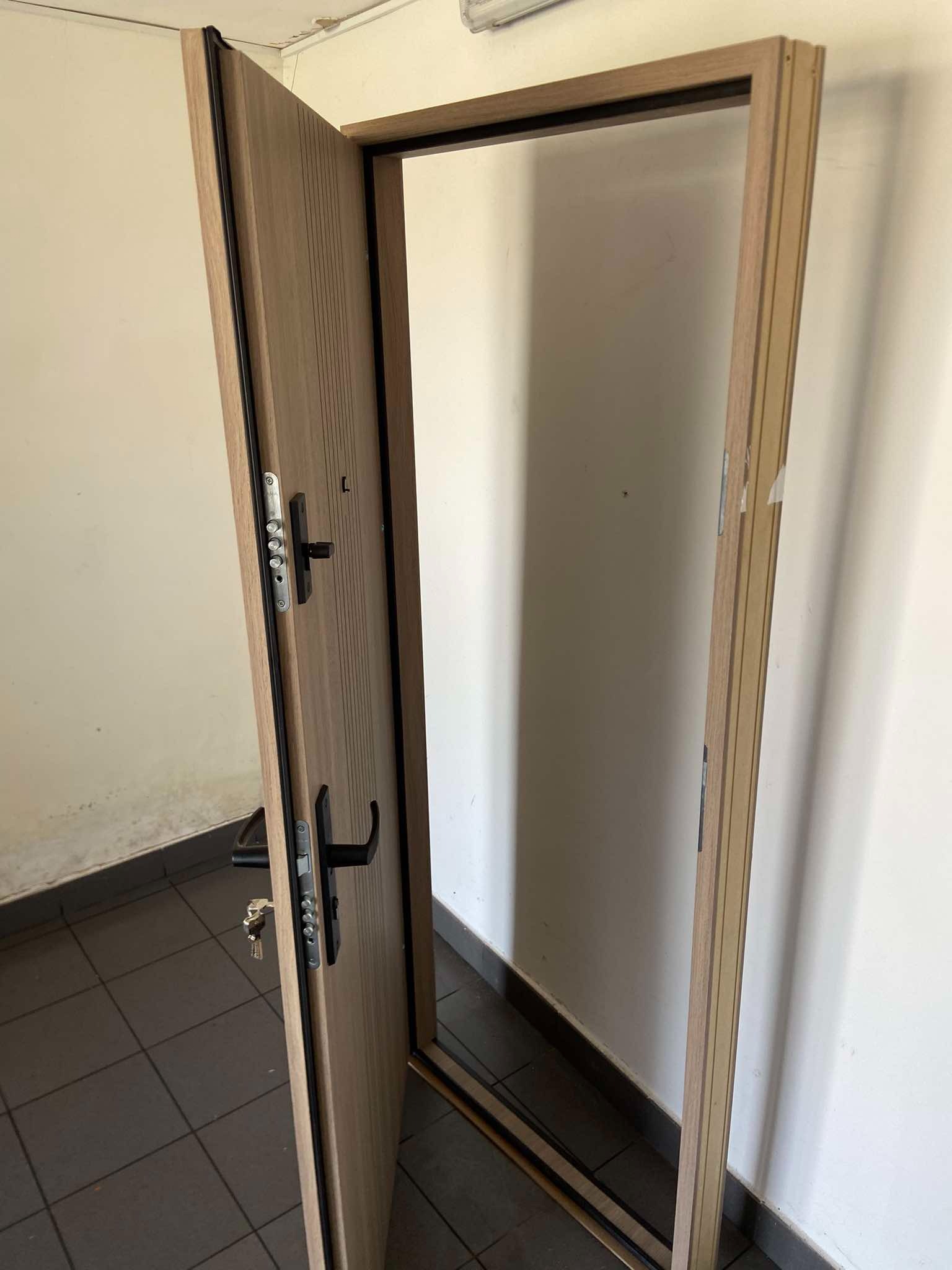 Otwarta, brązowa, ryflowana, zewnętrzna stolarka z widocznym zamkiem wielopunktowym i kluczami w klamce, osadzona w surowym otworze drzwiowym.