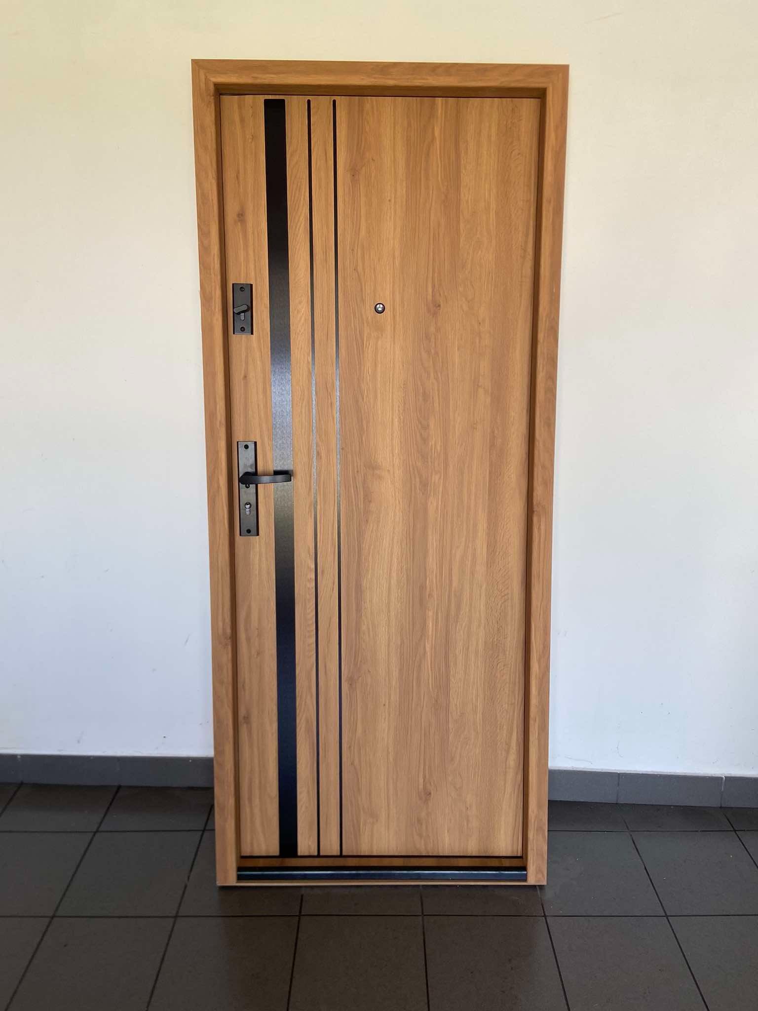 Drewniane drzwi wejściowe z czarnymi, pionowymi akcentami i metalową klamką na tle jasnej ściany i ciemnej podłogi z płytek.