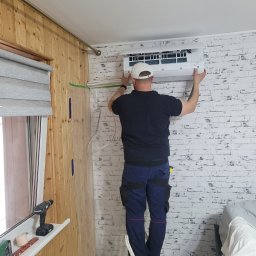 D&G EXPERT - Mężczyzna w białej czapce instaluje klimatyzator na ścianie z białej cegły w pokoju z drewnianą boazerią. Na parapecie leżą narzędzia.