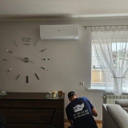 D&G EXPERT - Montaż klimatyzacji Haier w salonie: pracownik w firmowej koszulce D&G instaluje urządzenie na ścianie z dekoracyjnym zegarem. Widoczne narzędzia i meble.