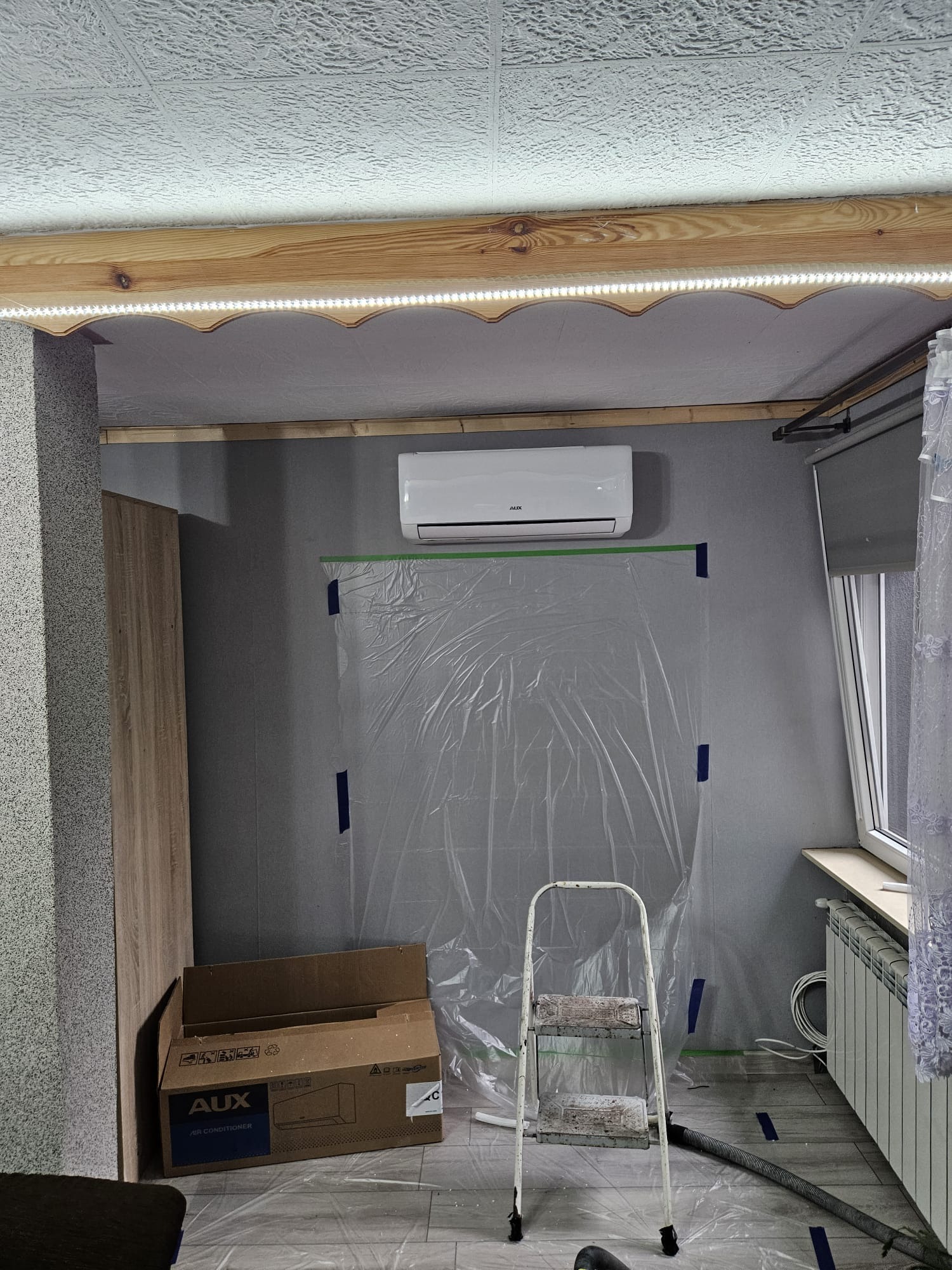 Biały klimatyzator marki AUX zamontowany na szarej ścianie, zabezpieczonej folią malarską, z widoczną skrzynią po urządzeniu i białą, metalową drabinką.
