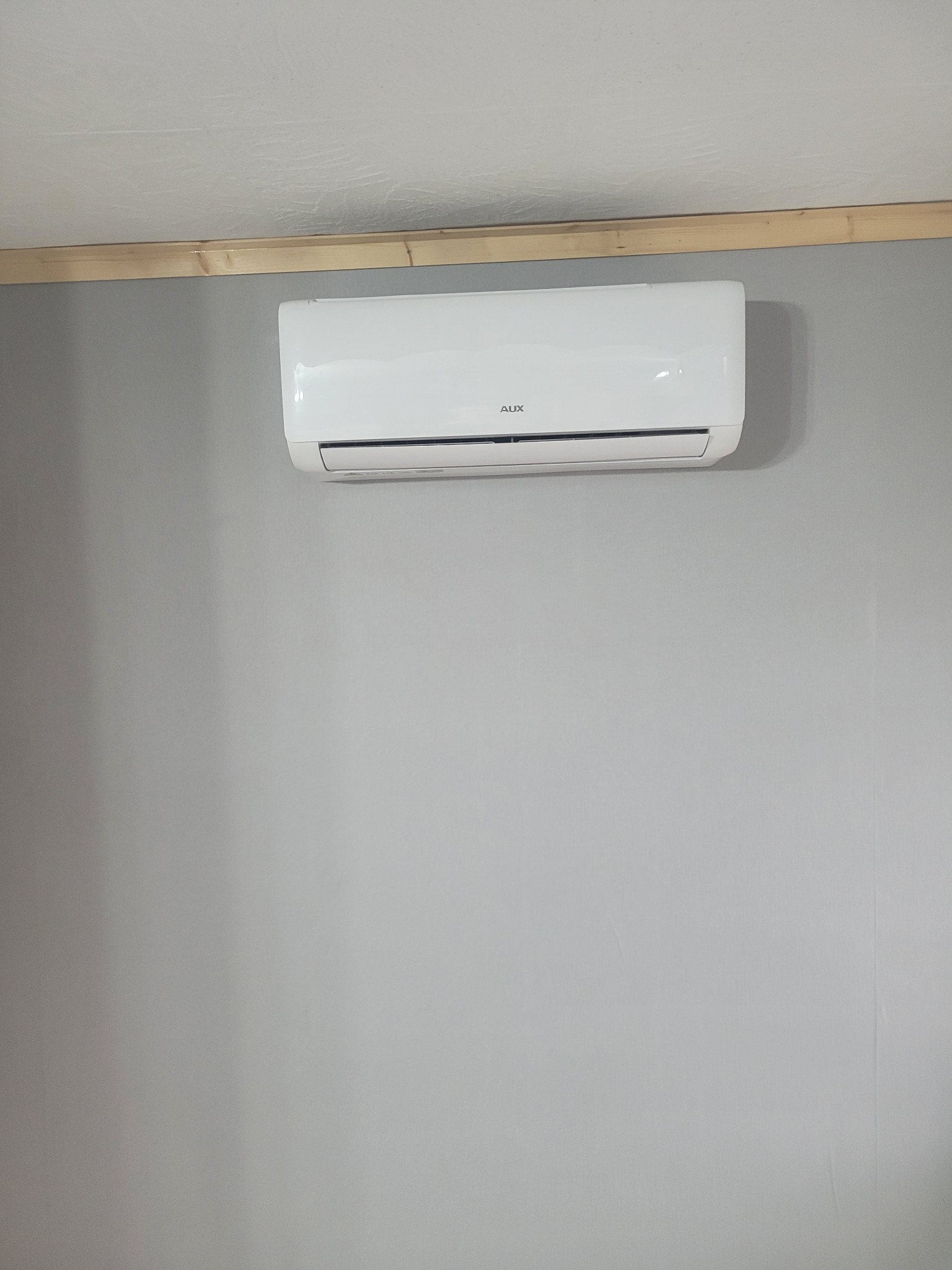Biała klimatyzacja AUX zamontowana na szarej ścianie w pomieszczeniu z drewnianą listwą podsufitową.