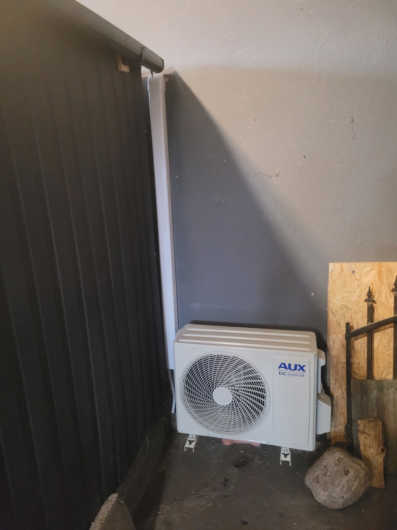 Biała jednostka zewnętrzna klimatyzacji AUX DC Inverter zamontowana przy szarej ścianie i czarnej blasze falistej, widoczny kamień na podłożu.