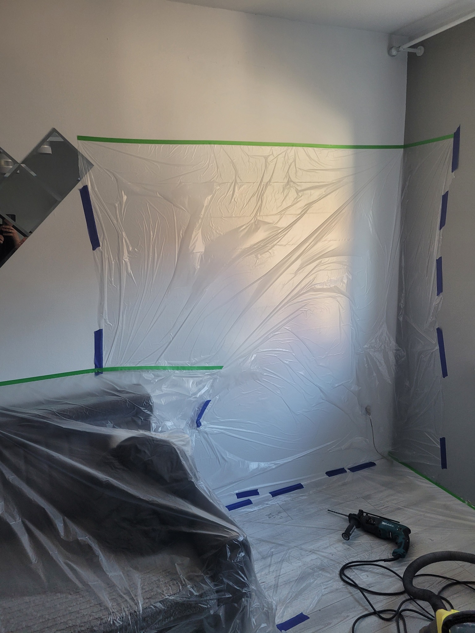 Przygotowanie pokoju do wiercenia otworów: ściany i meble zabezpieczone folią i taśmą malarską, wiertarka leży na podłodze.