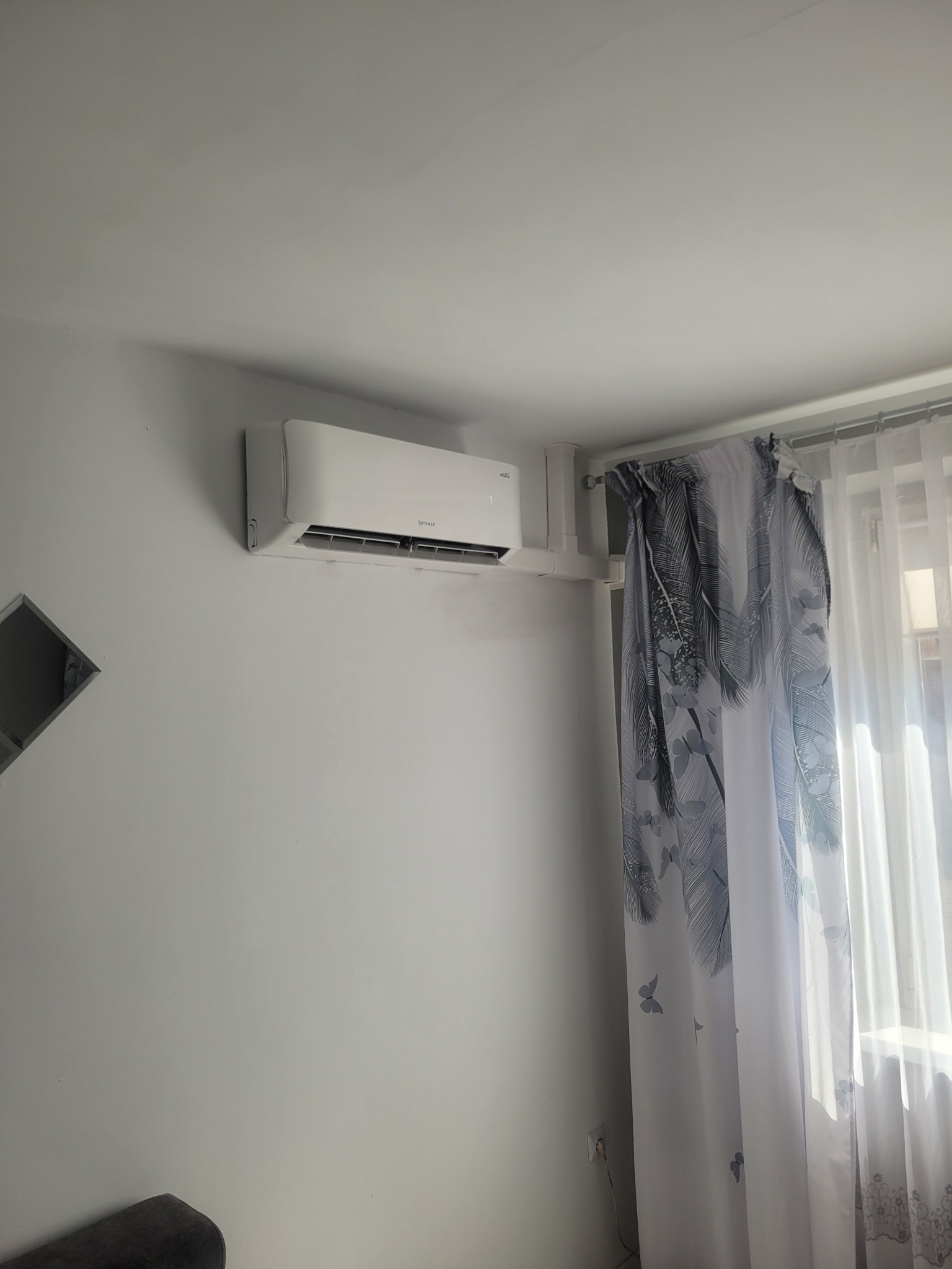 Biały klimatyzator marki Rotenso zamontowany na ścianie w pokoju, obok okna z zasłonami w pióra. Widoczny fragment sofy w rogu kadru.