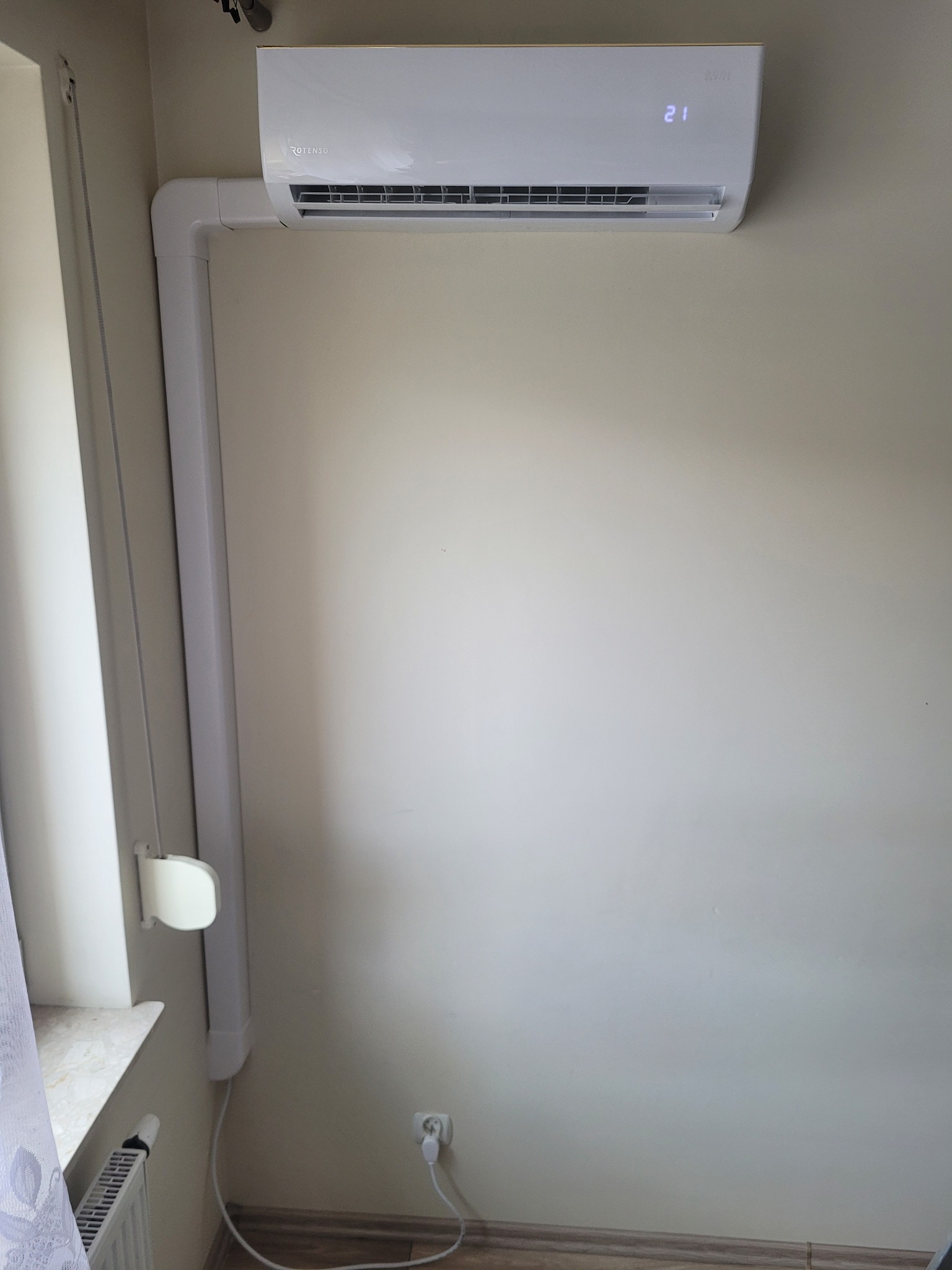 Biała klimatyzacja Rotenso na ścianie, z widoczną rurą odprowadzającą i wyświetlaczem pokazującym temperaturę 21 stopni. Widoczny fragment okna i grzejnika.