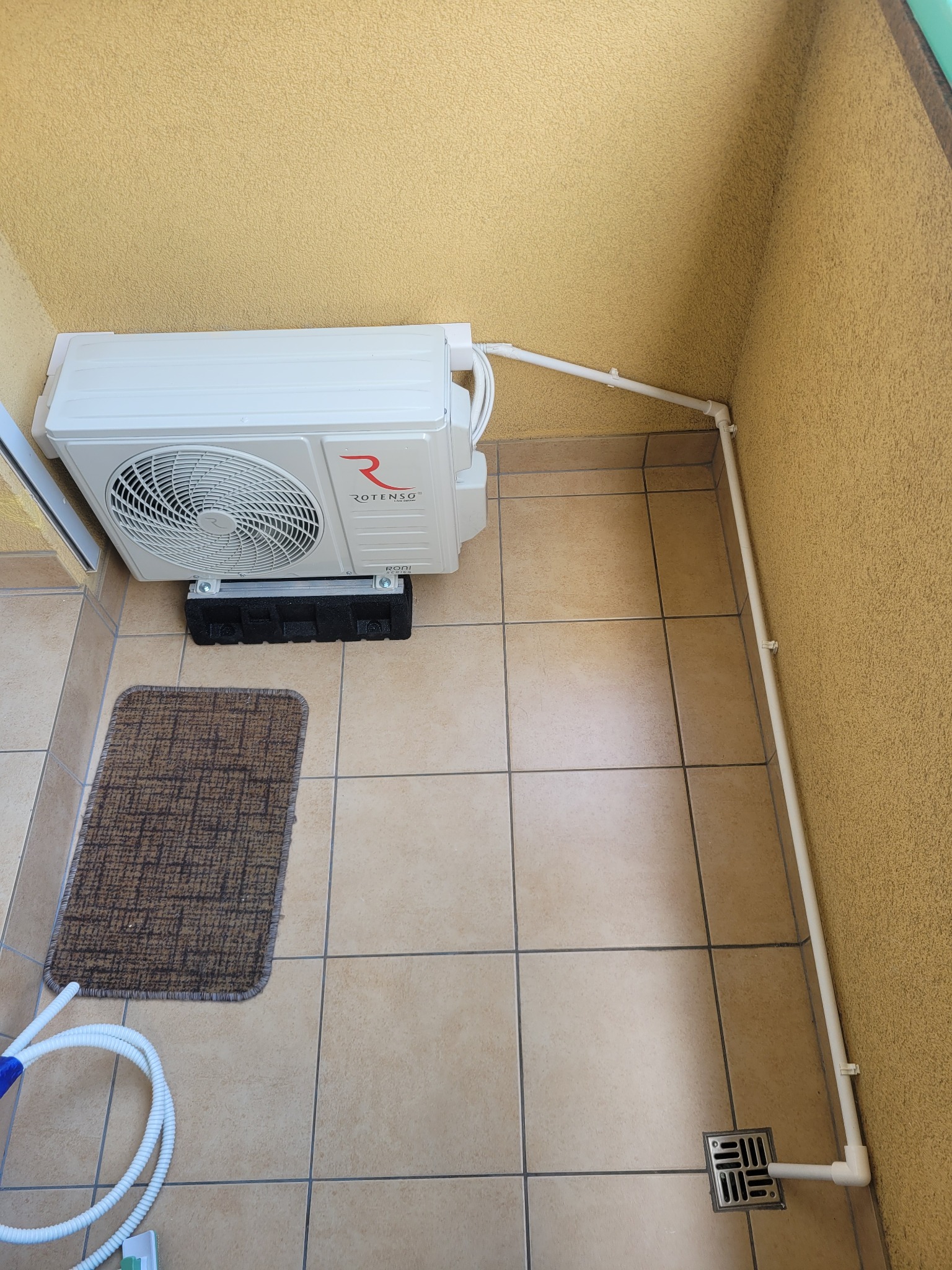 Zamontowany klimatyzator Rotenso na balkonie, z widoczną instalacją odprowadzającą skropliny. Ujęcie z góry, w tle żółta ściana o fakturze baranka.