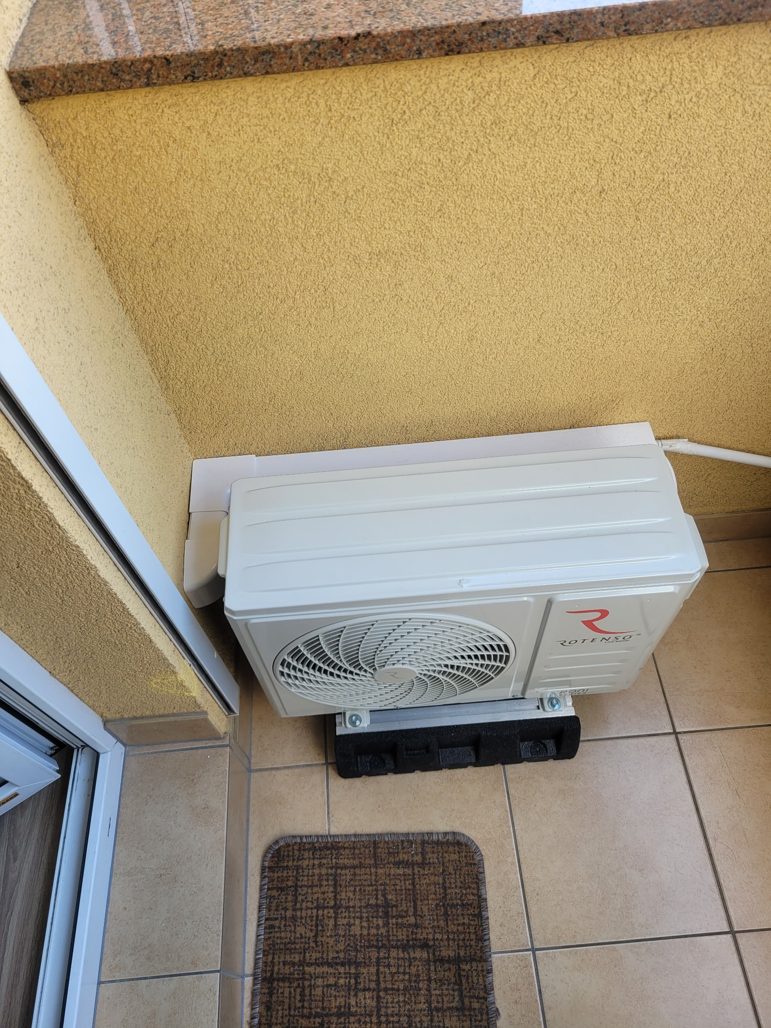 Biały klimatyzator Rotenso zamontowany na balkonie, widok z góry, z fragmentem drzwi balkonowych i wycieraczki. Żółta ściana z fakturą.
