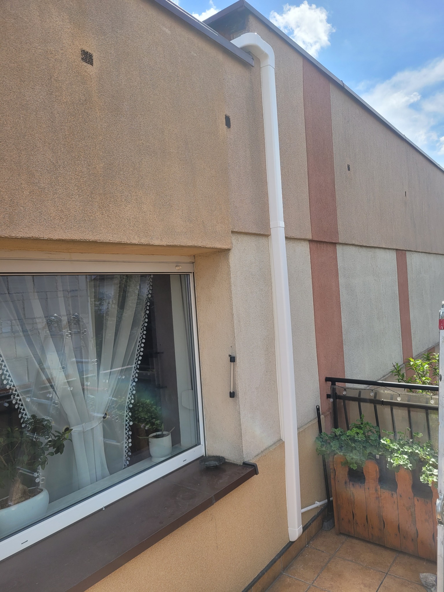 Biała rura klimatyzacji na elewacji budynku z oknem i balkonem, jasne tło nieba. Widoczne detale wykończenia i rośliny w donicach.