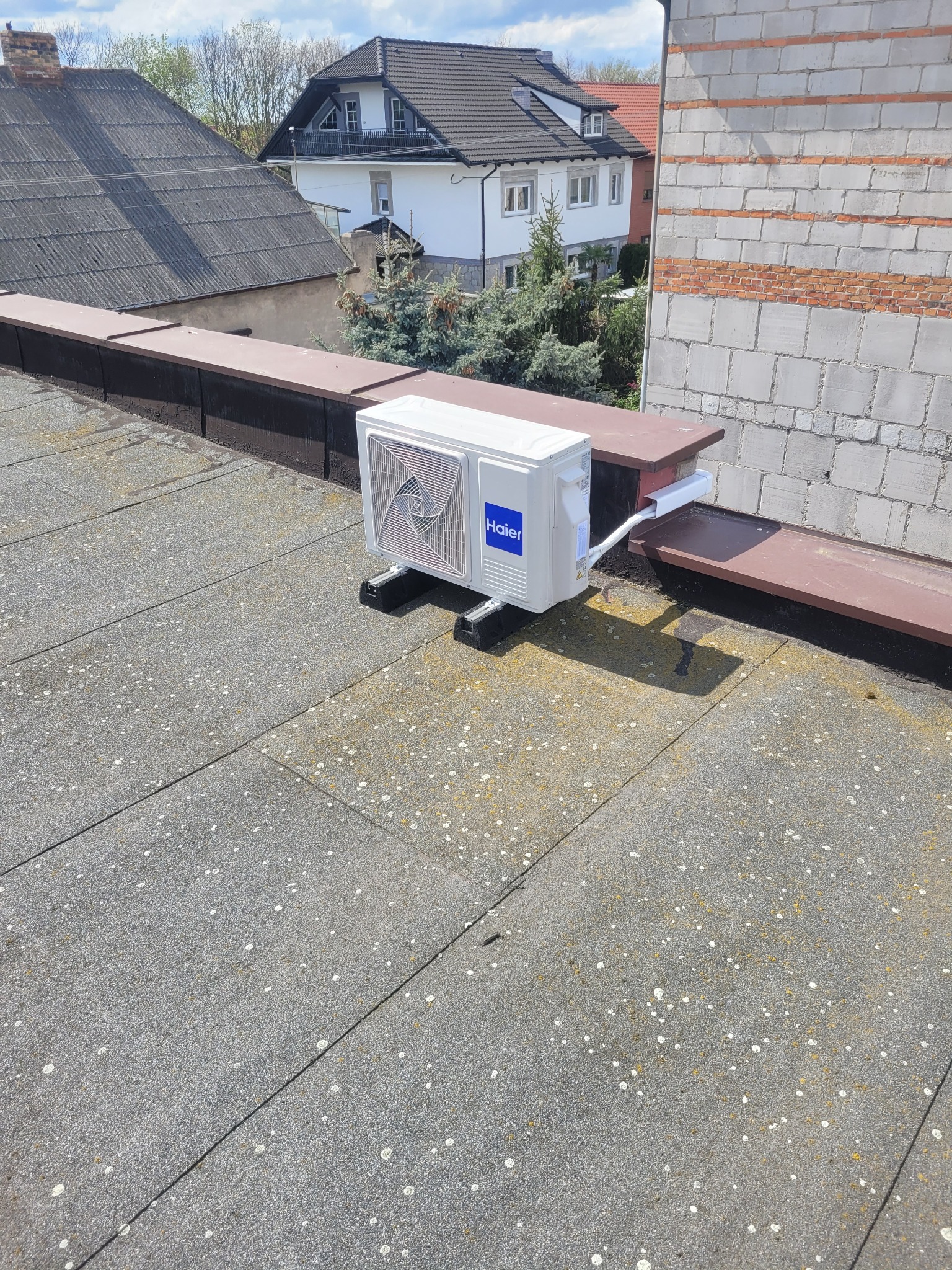 Biała jednostka zewnętrzna klimatyzacji Haier zamontowana na płaskim dachu budynku, widok z góry, z elementami instalacji i otoczeniem w tle.