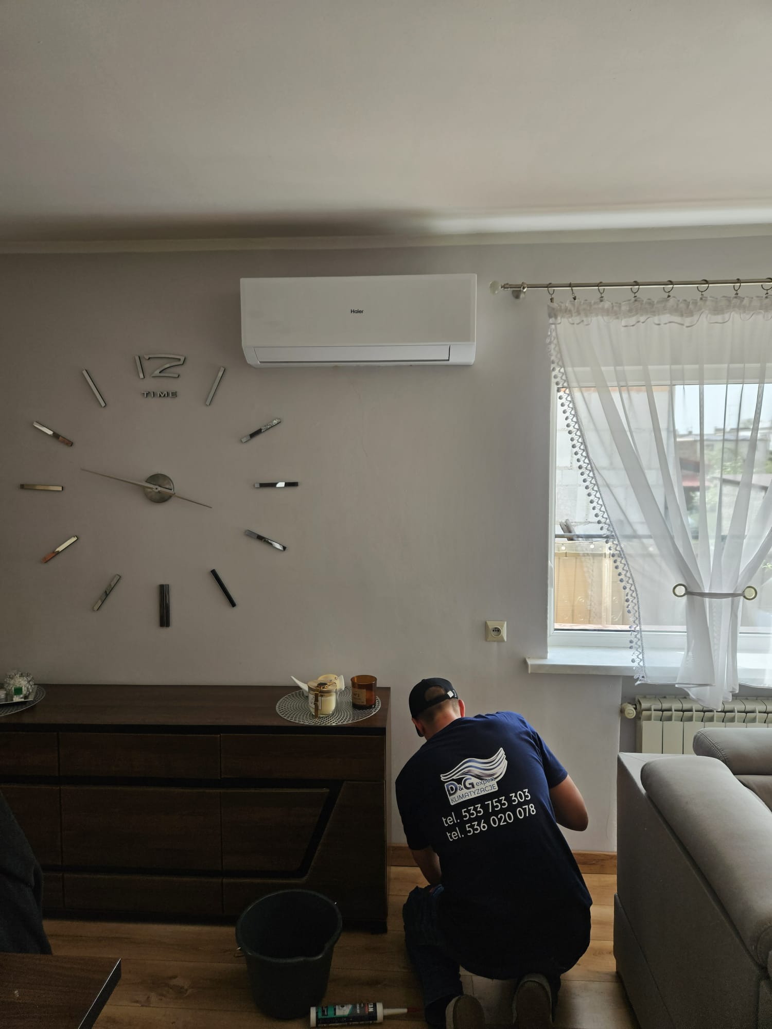 Montaż klimatyzacji Haier w salonie: pracownik w firmowej koszulce D&G instaluje urządzenie na ścianie z dekoracyjnym zegarem. Widoczne narzędzia i meble.