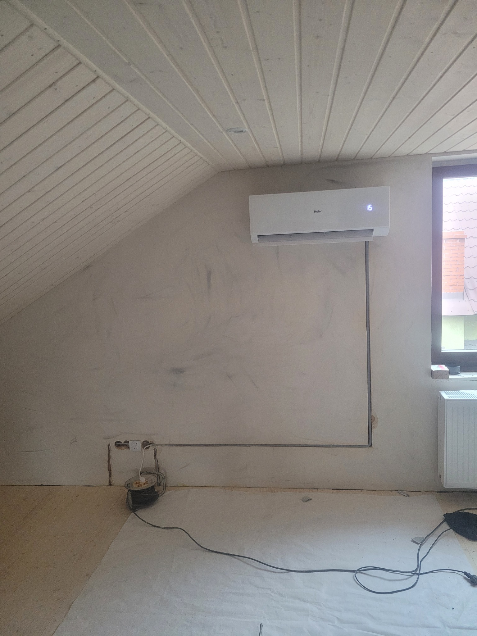 Biały klimatyzator Haier na ścianie w pokoju na poddaszu z boazerią. Widoczne przewody i gniazdko elektryczne. Etap instalacji klimatyzacji.