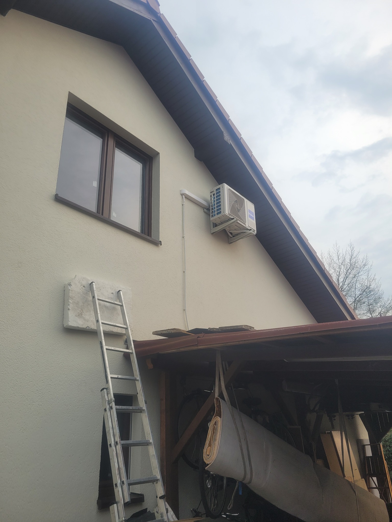 Biała jednostka zewnętrzna klimatyzacji Haier zamontowana na ścianie domu, z drabiną opartą o ścianę i fragmentem zadaszenia w tle.