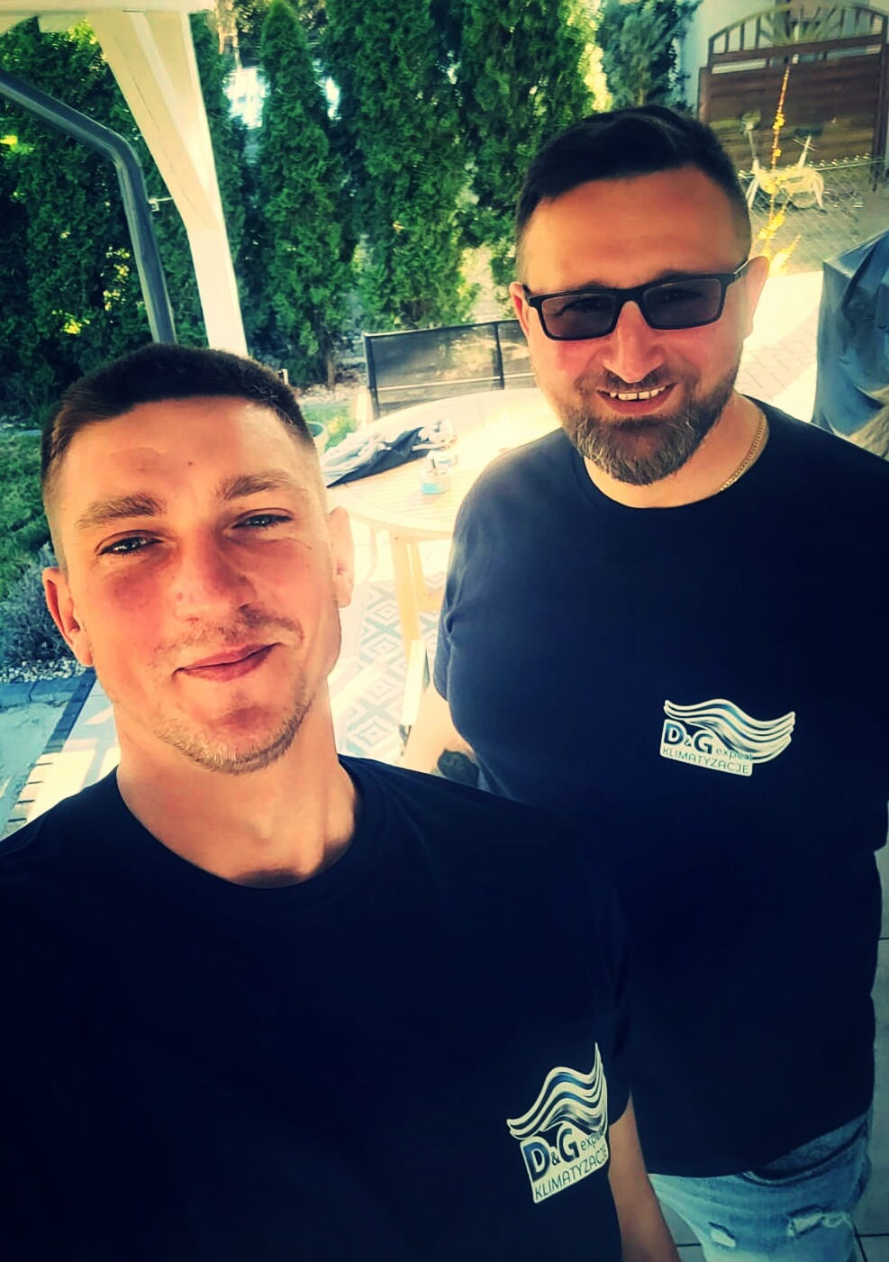 Uśmiechnięci mężczyźni w czarnych koszulkach z logo firmy klimatyzacyjnej na tle zieleni i białego stolika. Zdjęcie zrobione z bliska.